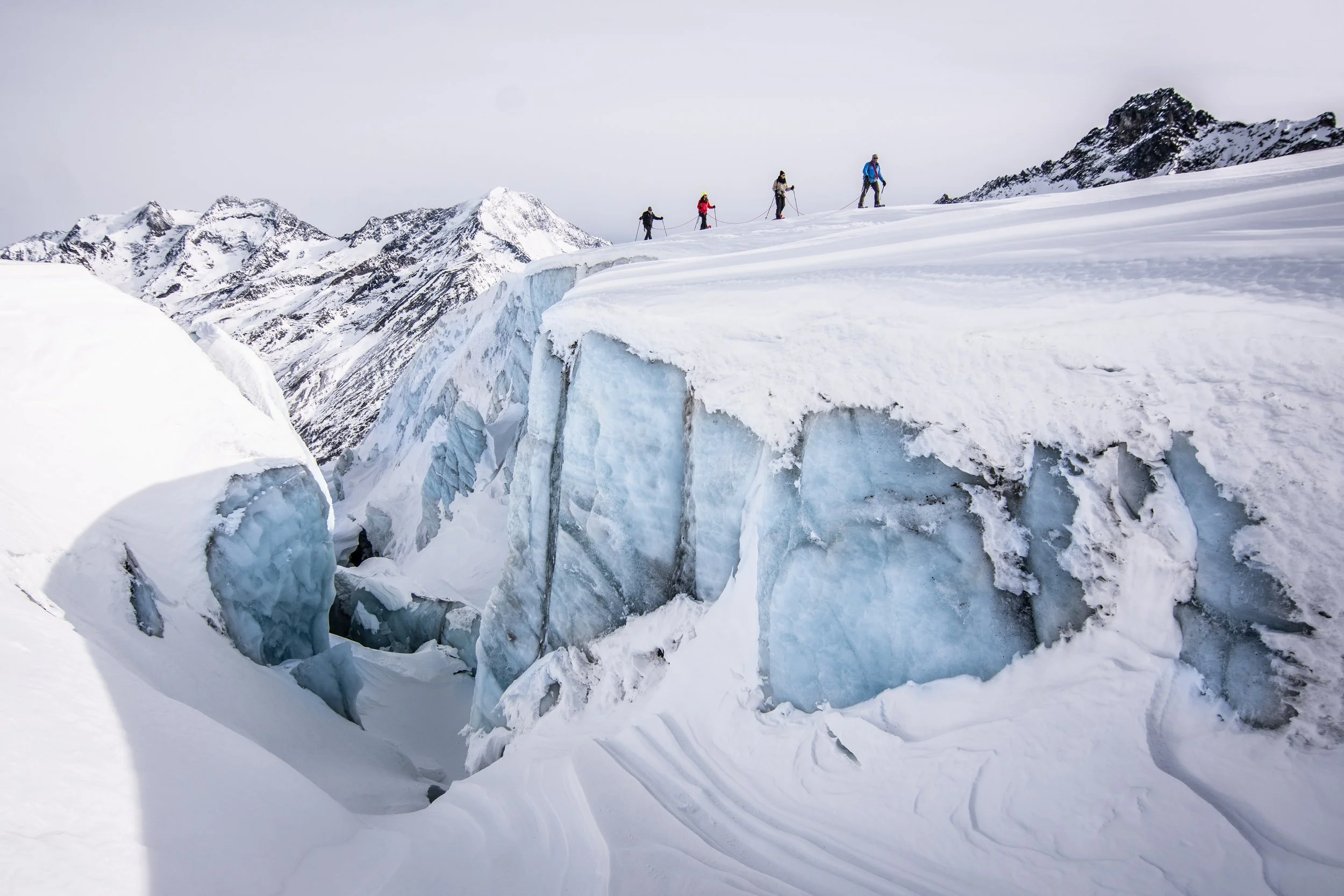 Group of hikers walking on snow-covered mountain terrain, with snow and ice formations and rugged mountain peaks in the background.