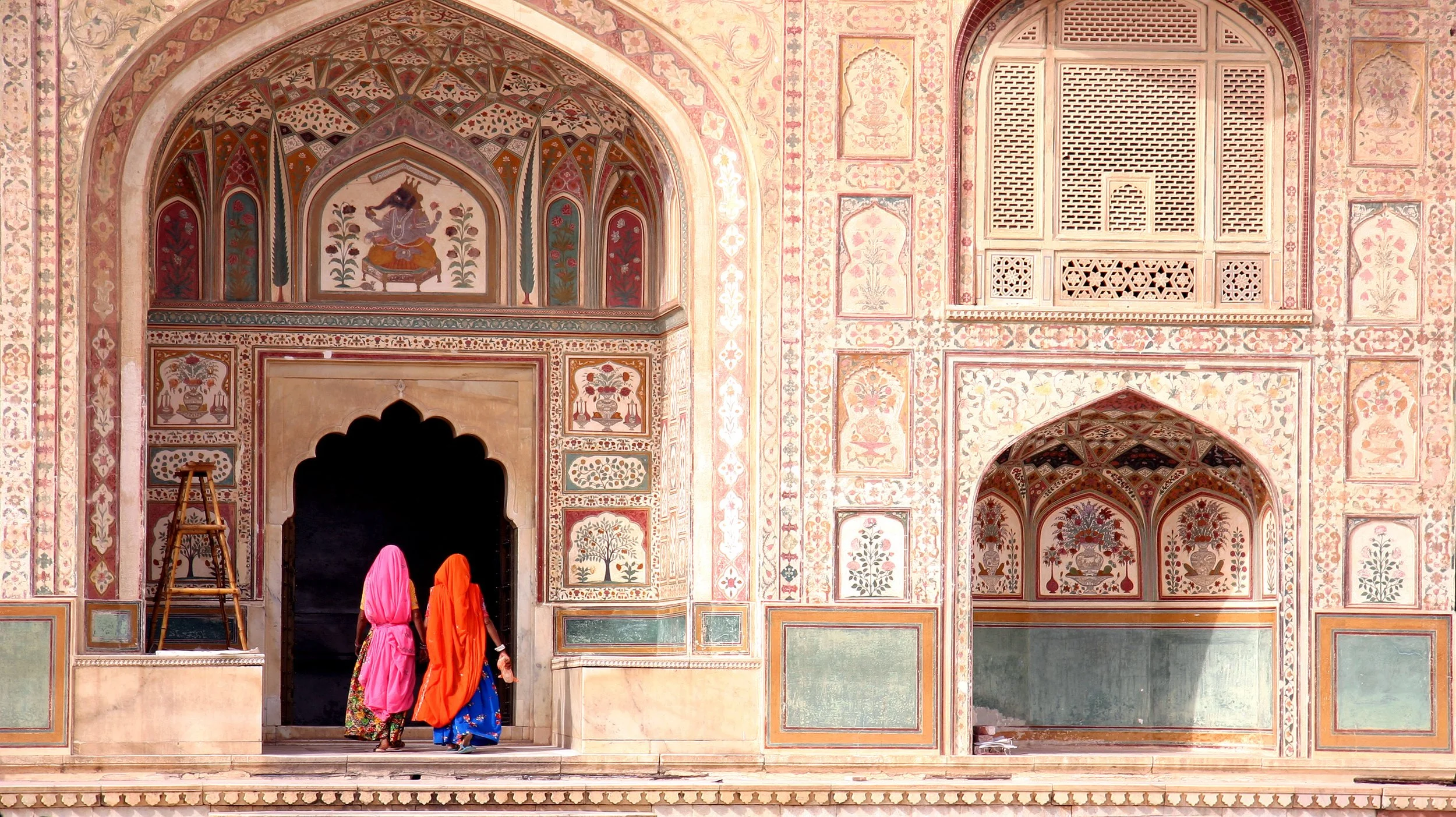 Three women dressed in vibrant traditional Indian clothing walking into an ornately decorated building with floral and geometric patterns.