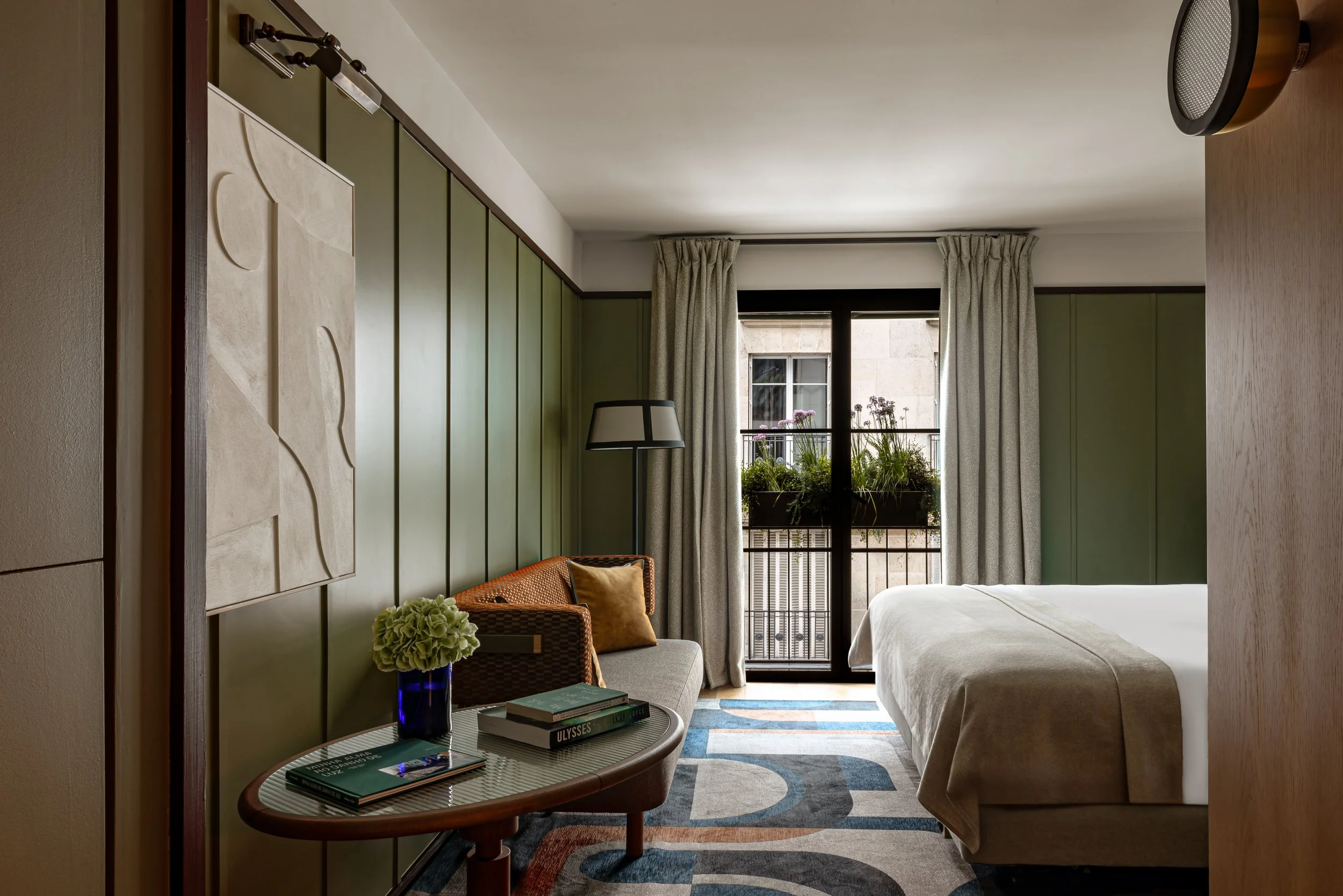 Hotel Norman Paris | Hotel room with green paneled wall, white bed, window with curtains, small table with books and a blue vase with flowers, wicker chair with yellow pillow, and patterned rug.