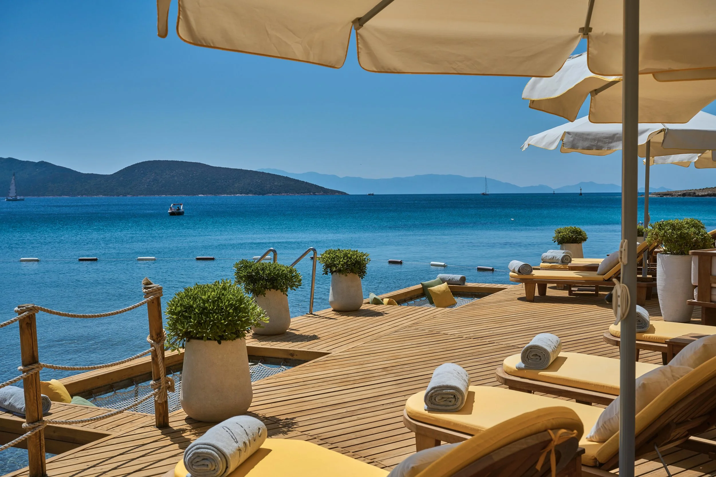 Beachside seating area with yellow lounge chairs, rolled towels, potted plants, large white umbrellas, overlooking blue water, with mountains and sailboats in the distance.