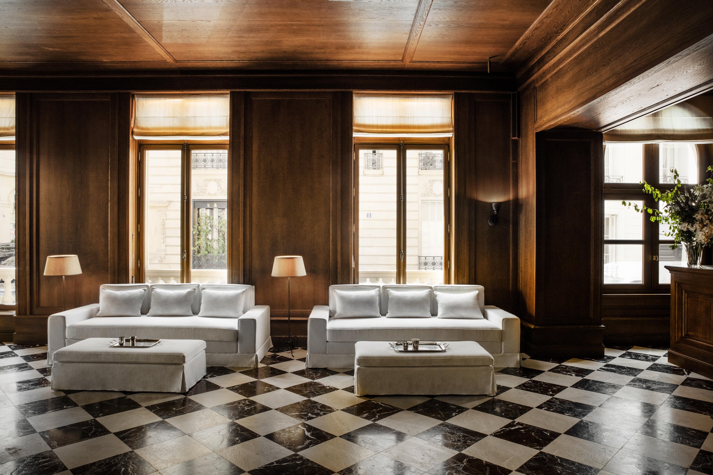 Hotel Balzac Paris | Elegant hotel or lounge area with white sofas, checkered black and white marble floor, large windows with roman shades, and dark wood paneled walls.