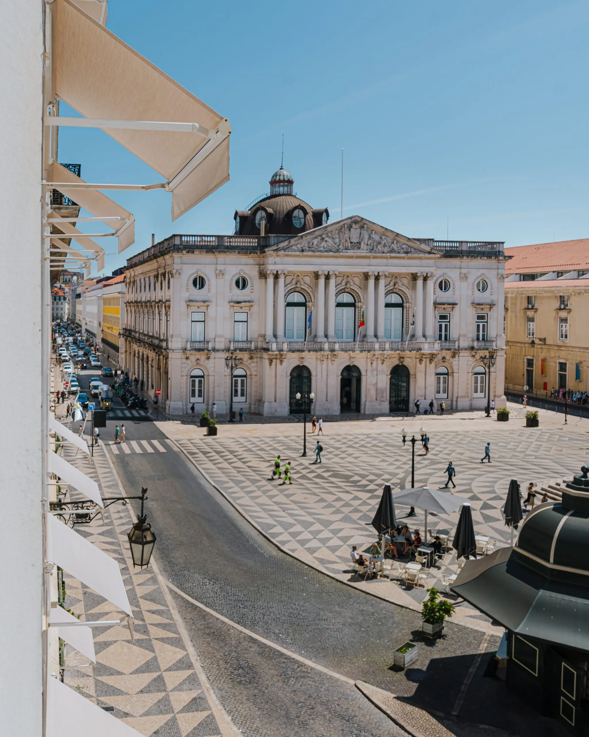 A city square with a patterned pavement, outdoor seating under umbrellas, and historic buildings including a prominent building with a dome, columns, and flags.