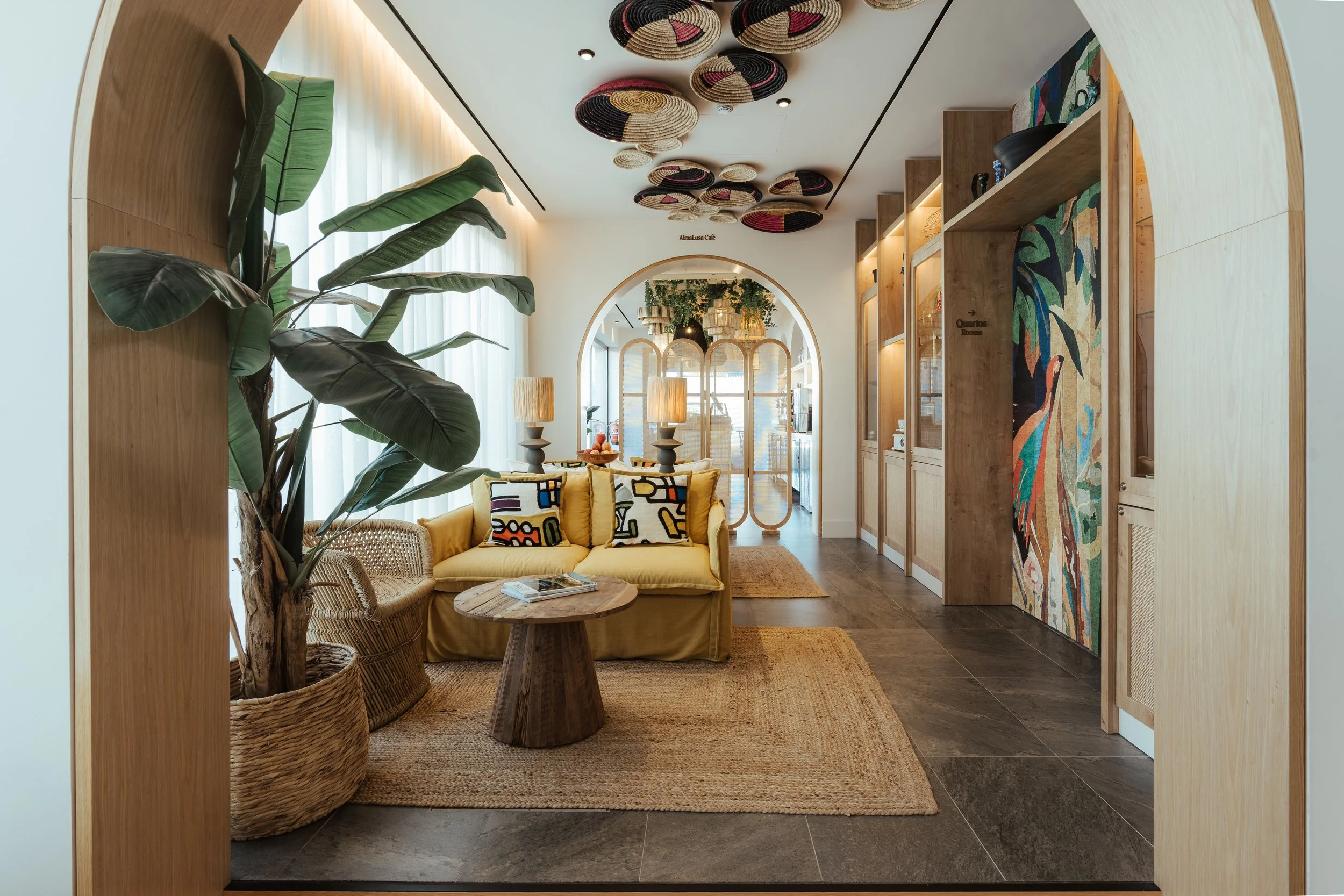 A cozy living room with a mustard yellow couch, decorative pillows, a wooden coffee table, a large potted plant, and wall art. The room features natural wood finishes, colorful woven ceiling decorations, and soft natural light streaming through sheer curtains.