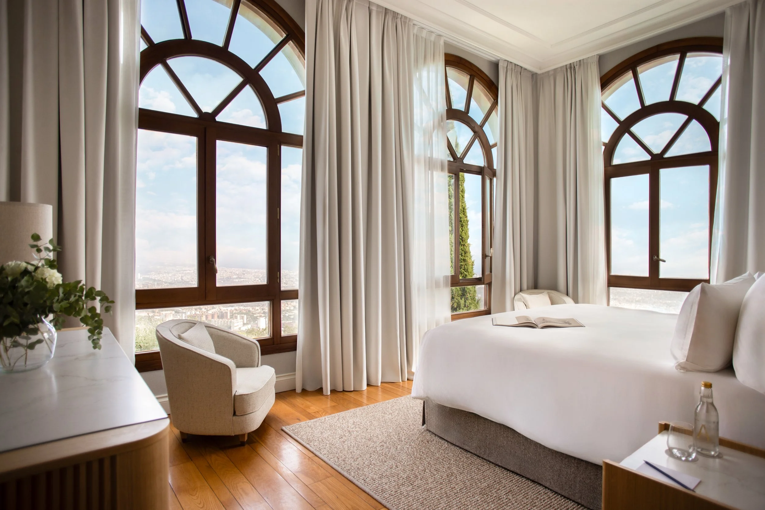 A hotel room with large arched windows, white curtains, and a view of the city and sky outside. There is a bed with an open book on it, a chair, and a small table with a plant and decorative items.
