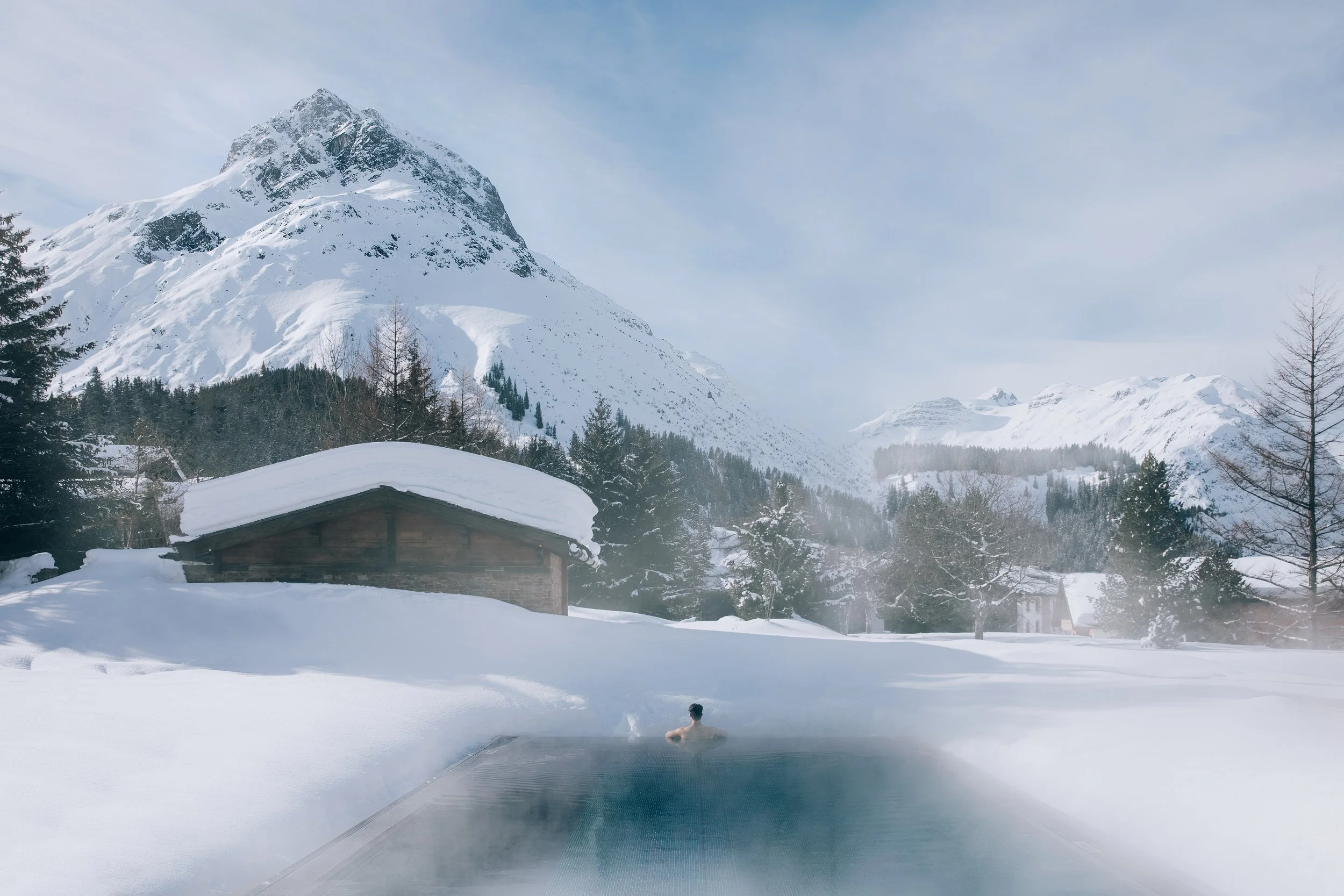 A hot spring pool with a person relaxing in the hot water, surrounded by snow-covered mountains and trees, with a cloudy sky overhead.