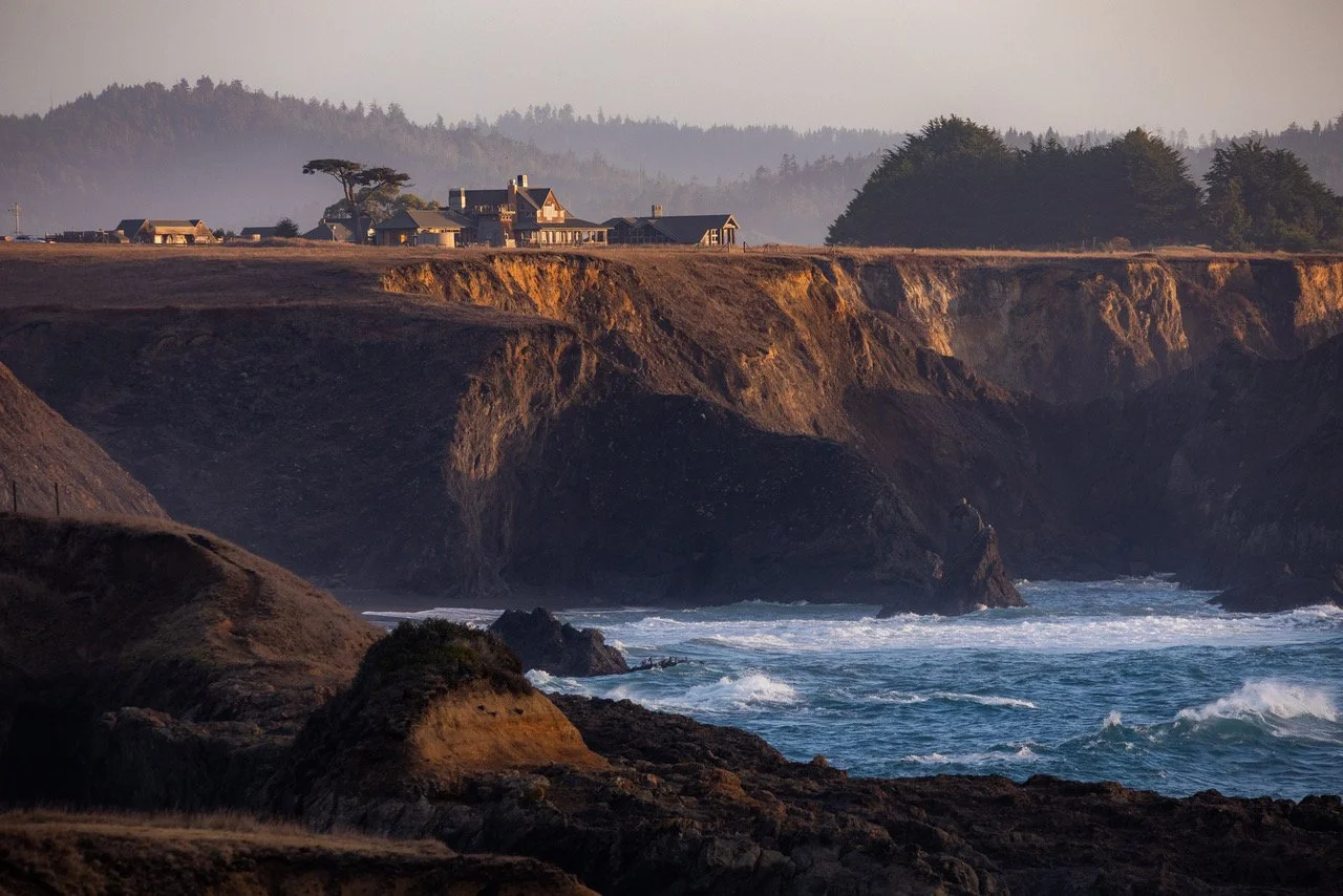 Cliffside coastal landscape during sunset with rugged cliffs, ocean waves, and houses on the hilltop.