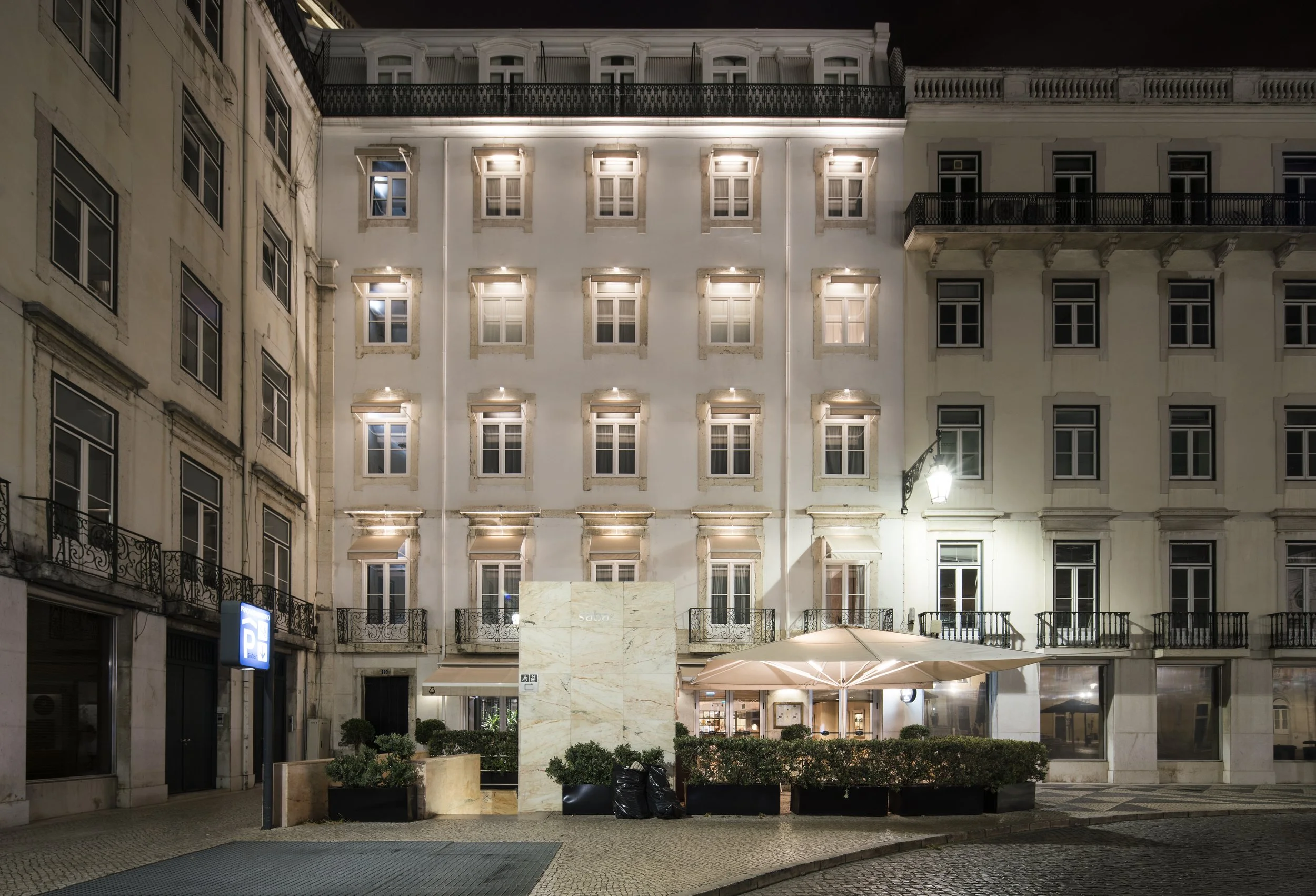 Nighttime view of a white building with multiple illuminated windows and balconies, with some greenery and umbrellas at street level.