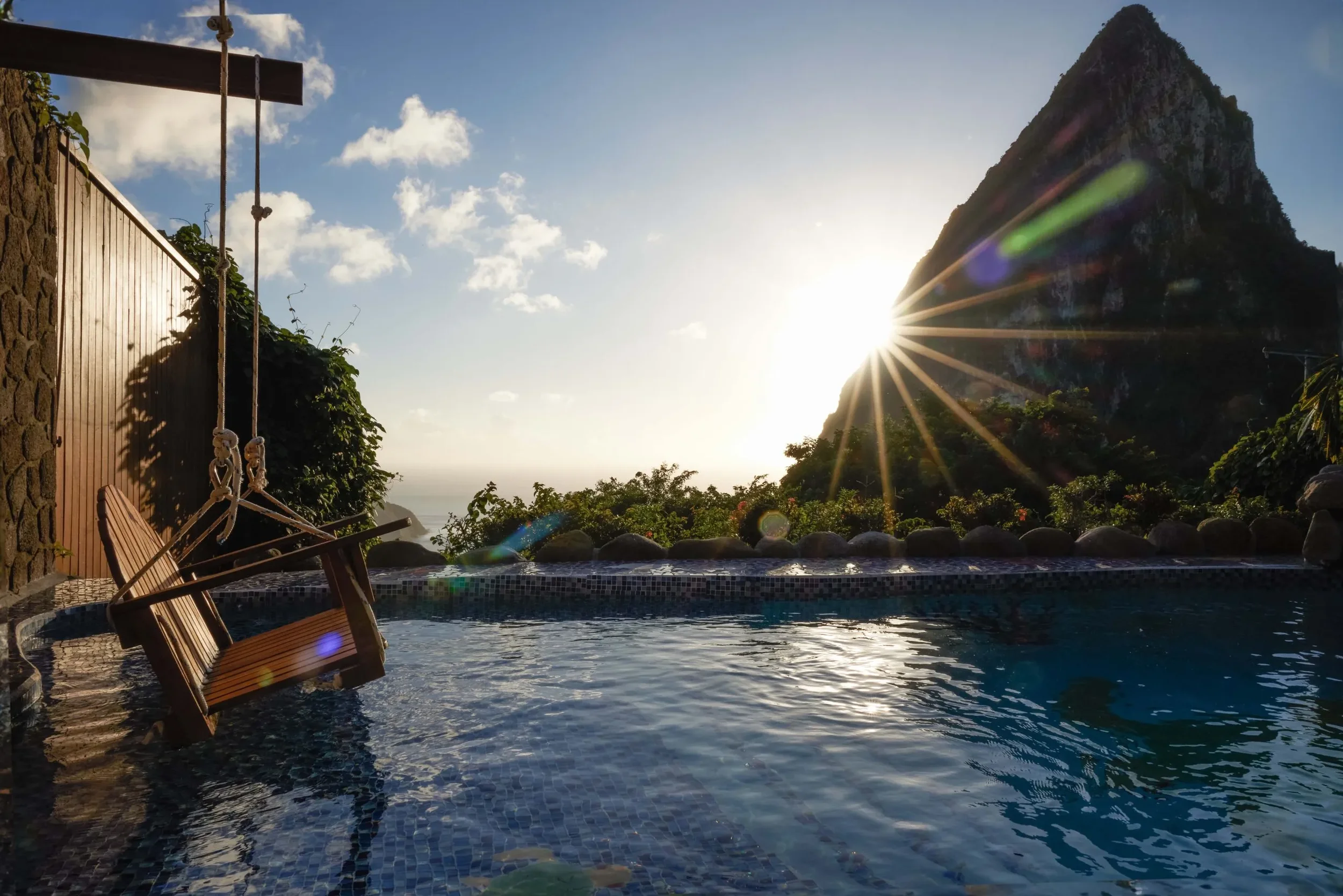 A swimming pool with a wooden lounge chair and a bench on the edge, surrounded by greenery and rocks, with a mountain in the background and the sun setting behind it, creating lens flare effects.