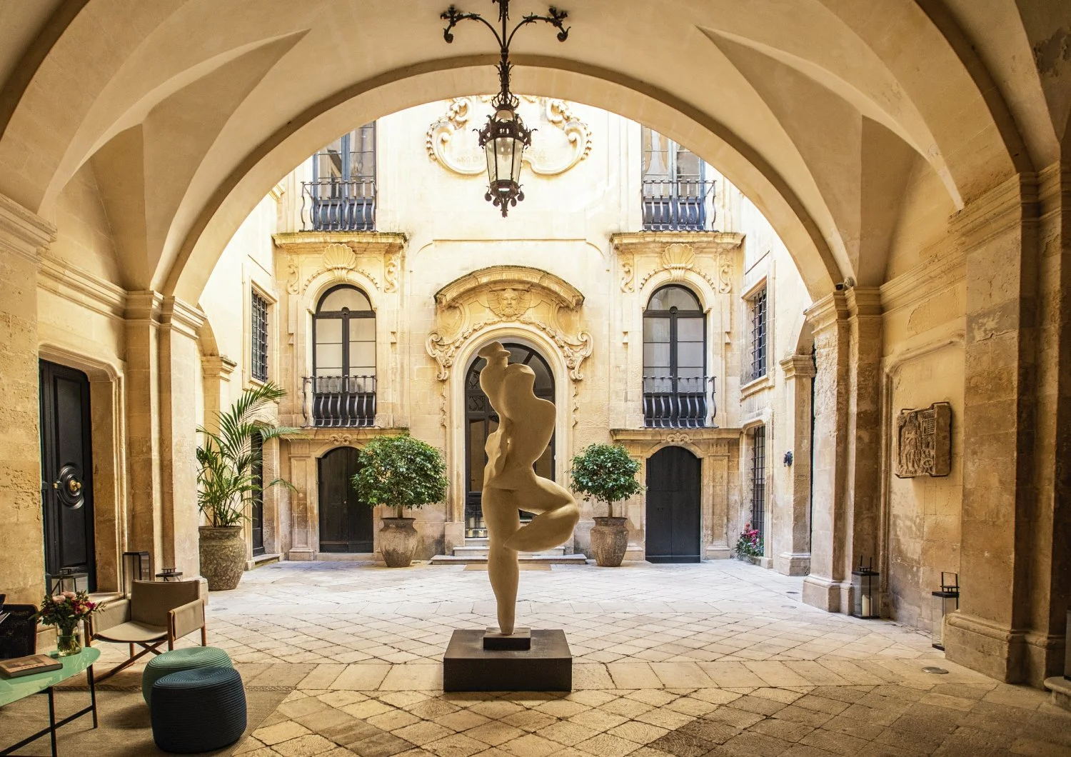 A courtyard with a classical sculpture of a woman in a pose, surrounded by potted trees and decorated with ornate architecture, including arches and balconies.