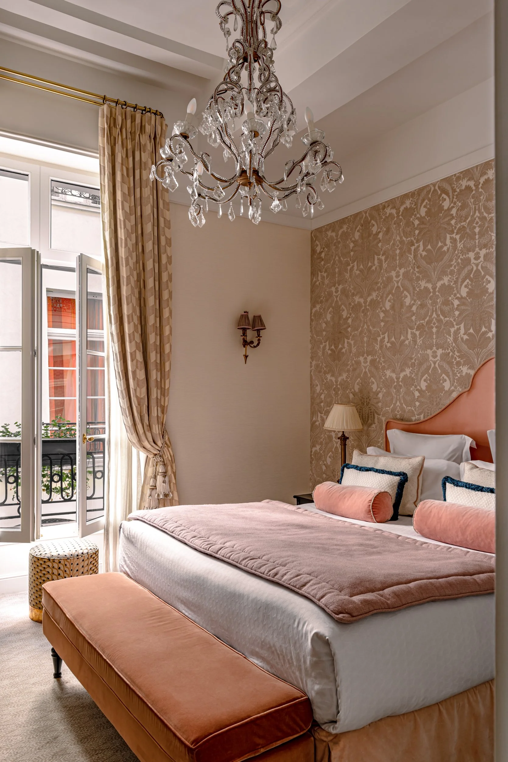 This image shows a luxurious bedroom with a pink bed and matching pink cushioned bench at the foot of the bed. There are pink cylindrical pillows on the bed, along with two decorative pillows with dark blue trim. A small bedside table with a beige lampshade and a wall sconce are visible. The room has a large window with patterned gold curtains, and a chandelier hangs from the ceiling.
