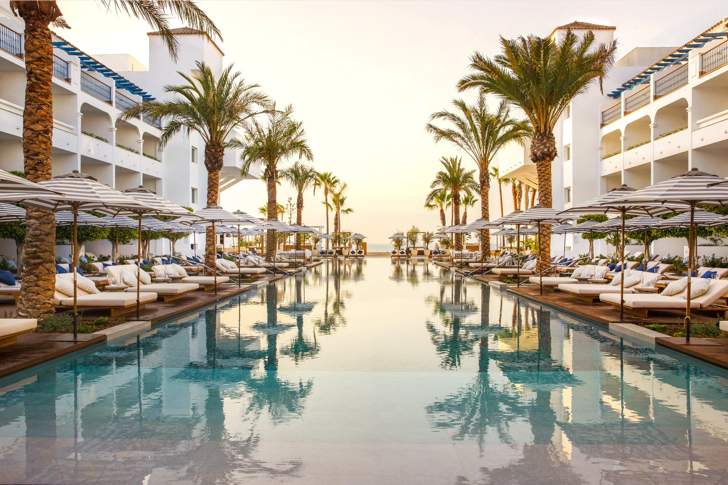 A luxurious hotel pool area with palm trees, white cabanas with cushions, and striped umbrellas. The pool reflects the sky and surrounding buildings with balconies, suggesting a tropical or coastal resort setting.