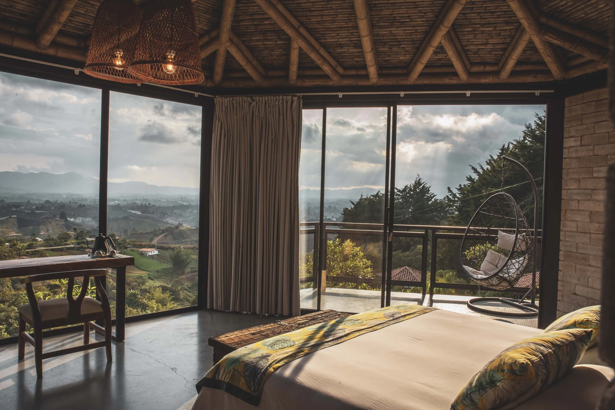 A cozy room with a bed, a hanging chair, and a view of mountains and countryside through large glass doors.