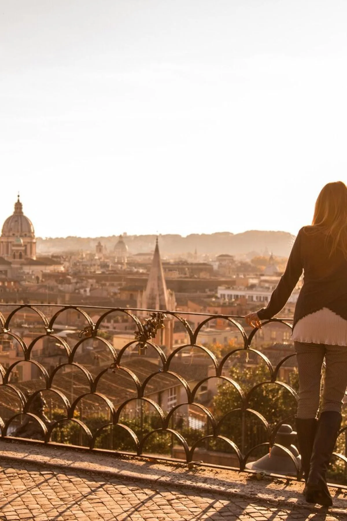 A woman looking over a cityscape from a balcony with a metal railing, possibly in Rome, Italy, during sunset.