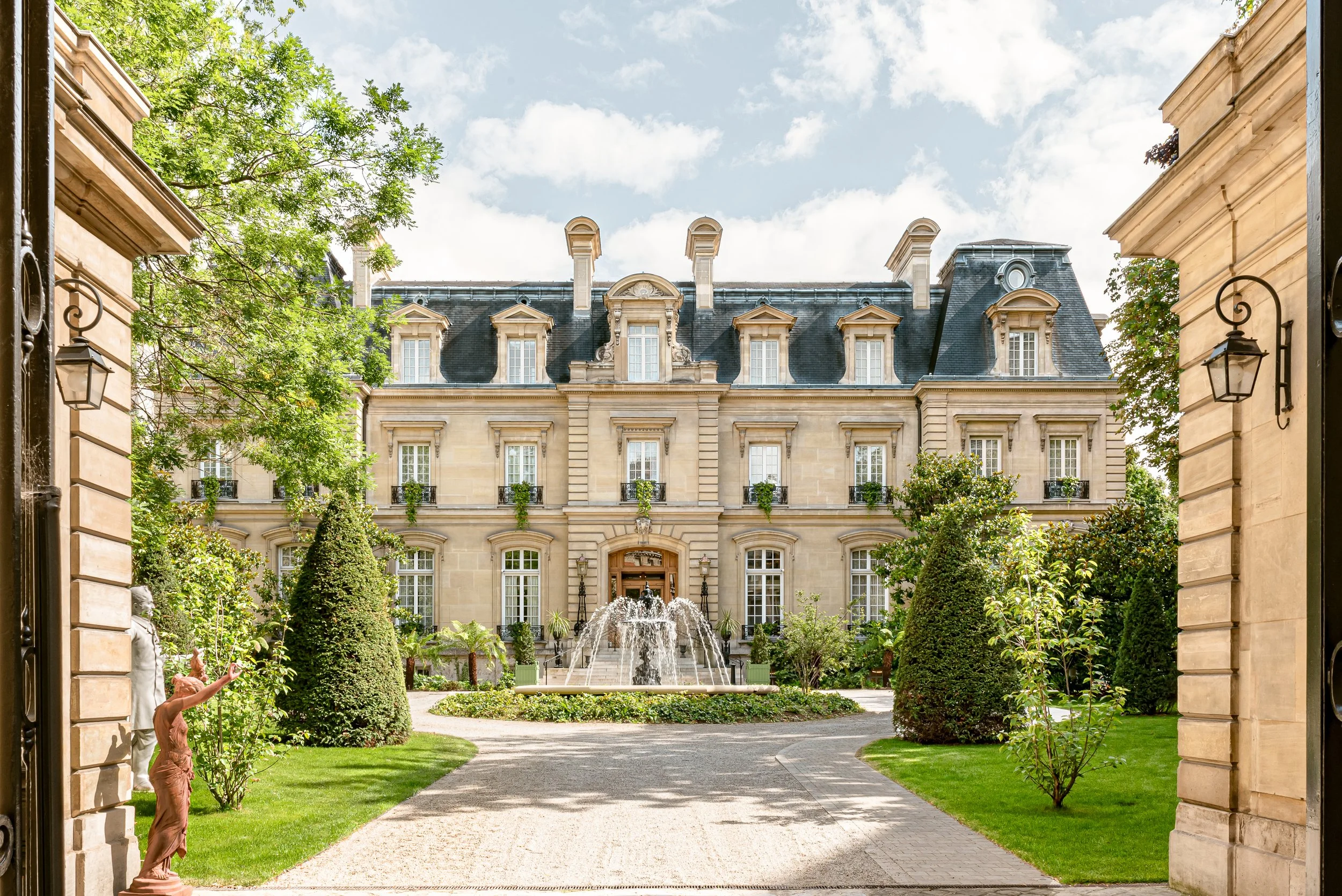 A view of a grand European-style building with a fountain in front, seen through an open gate, surrounded by well-manicured gardens and trees, under a partly cloudy sky.