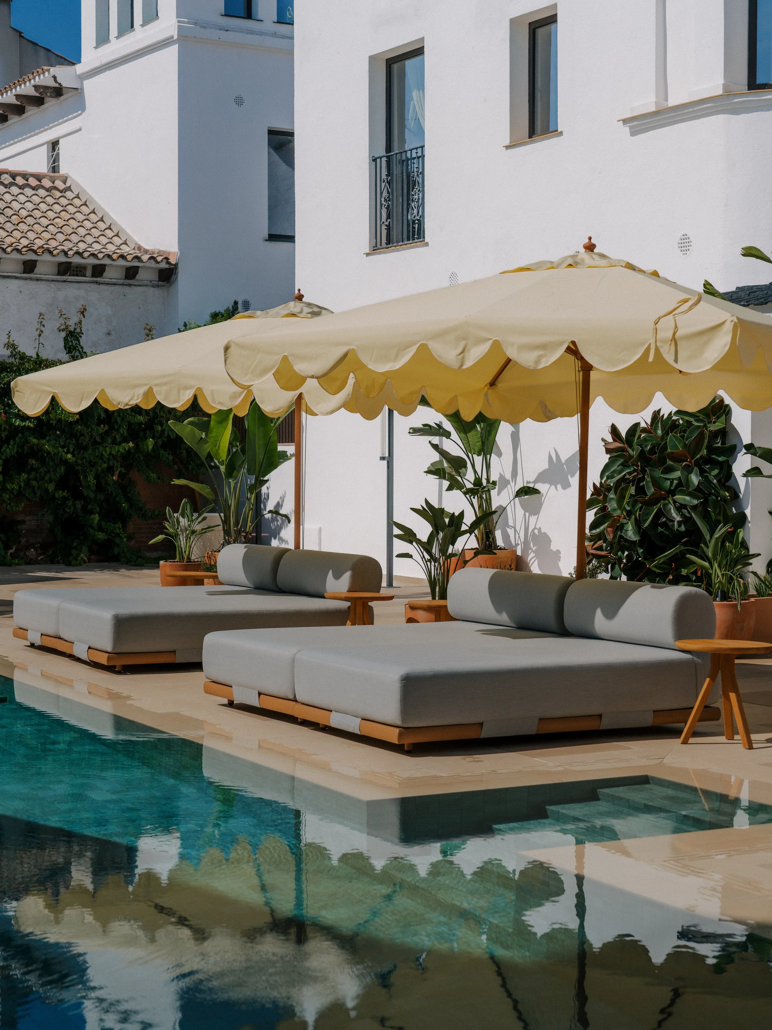 Modern outdoor poolside lounge area with white cushioned seating, yellow canopy umbrellas, and potted plants against a white building with windows and balconies.