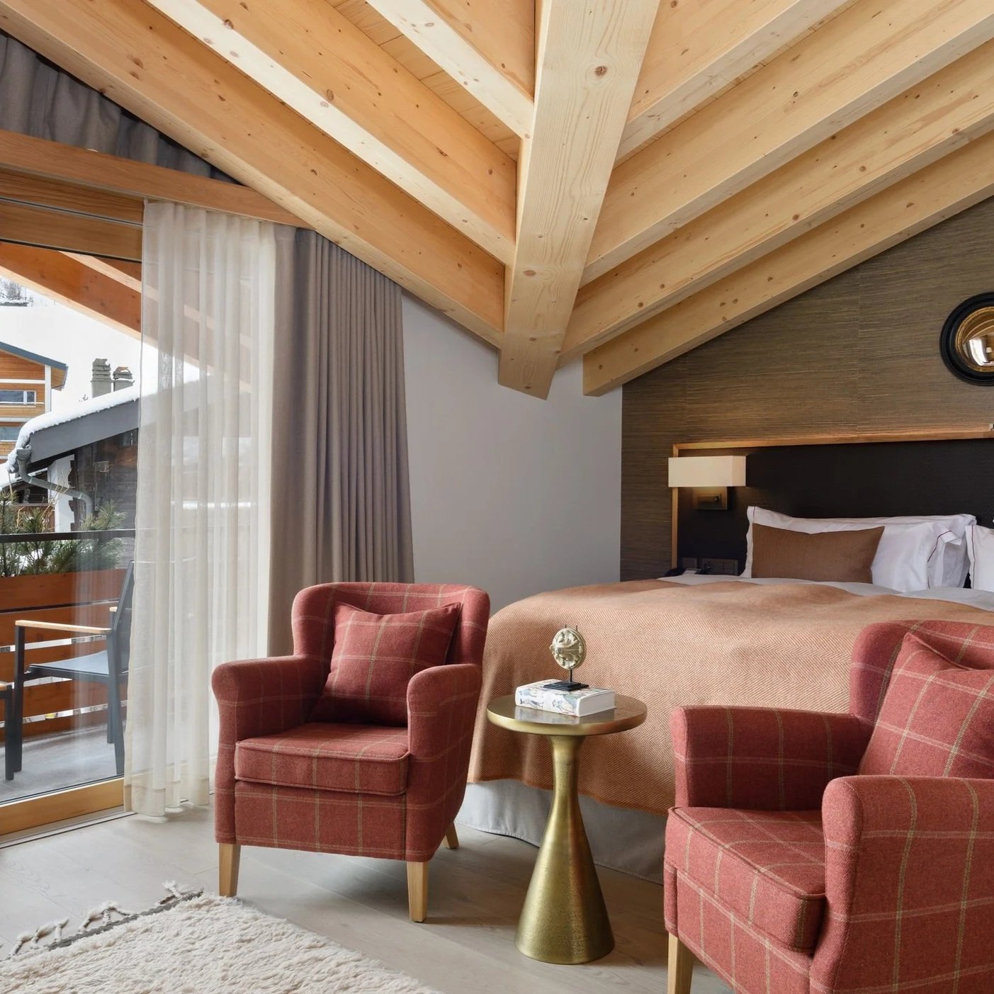 Hotel room with a bed, two red plaid armchairs, a small gold side table with a clock and books, a large window with white and gray curtains, and wooden ceiling beams