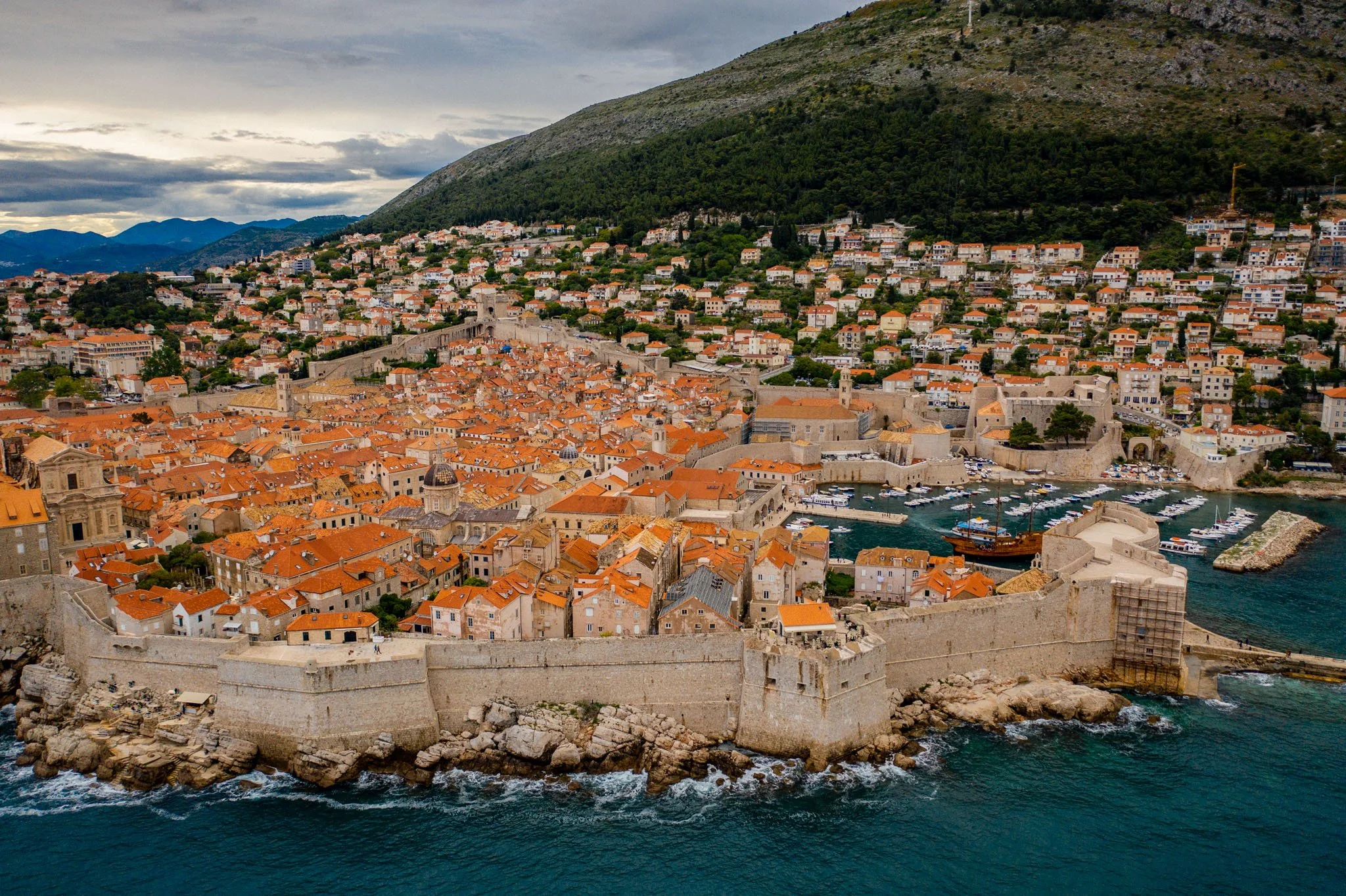 Aerial view of a historic coastal city with orange-tiled rooftops, stone walls, and a harbor filled with boats, surrounded by green hills and mountains under a cloudy sky.