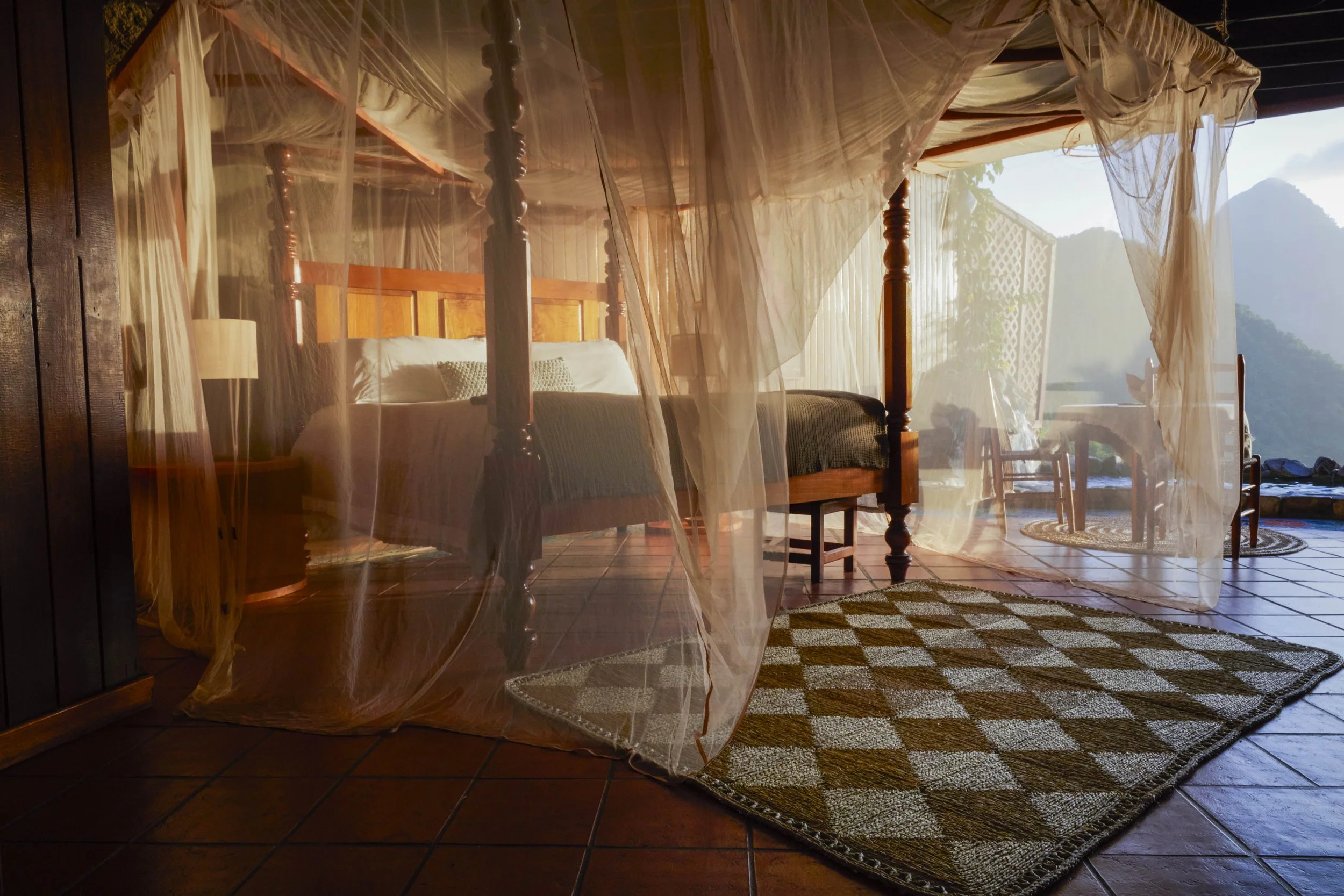A bedroom with a four-poster bed with sheer curtains, a bedside lamp, a tiled floor, a patterned rug, and an outdoor view of mountains and greenery in the distance.