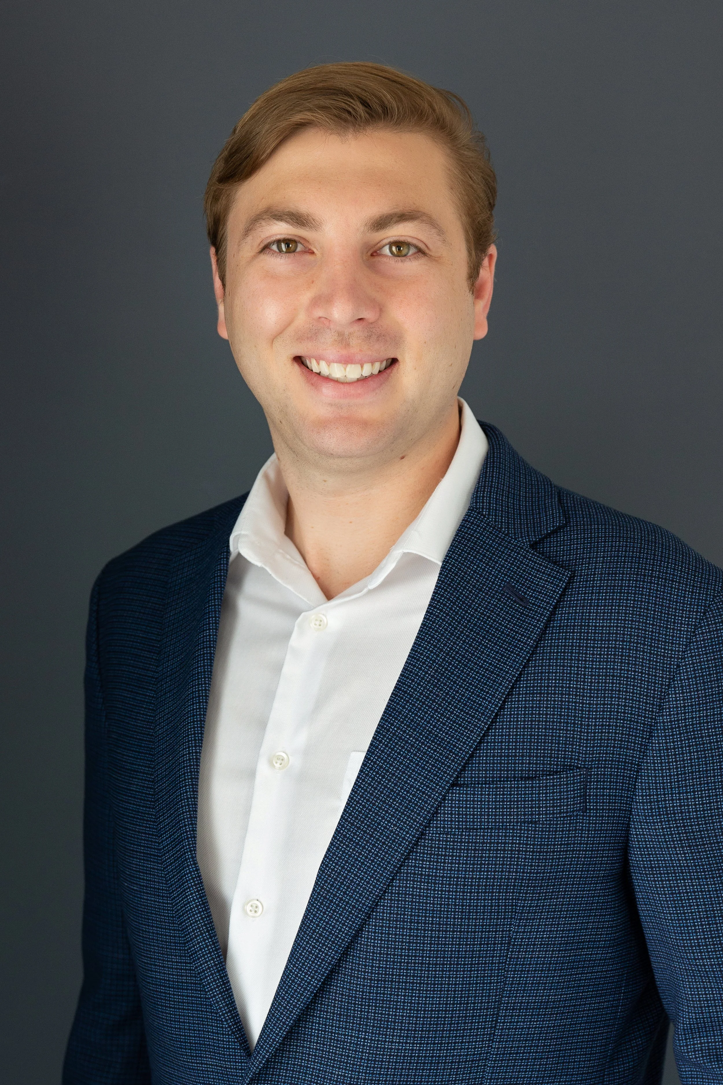Young man with light brown hair and hazel eyes, wearing a white dress shirt and a dark blue blazer with a small checkered pattern, smiling against a dark gray background.