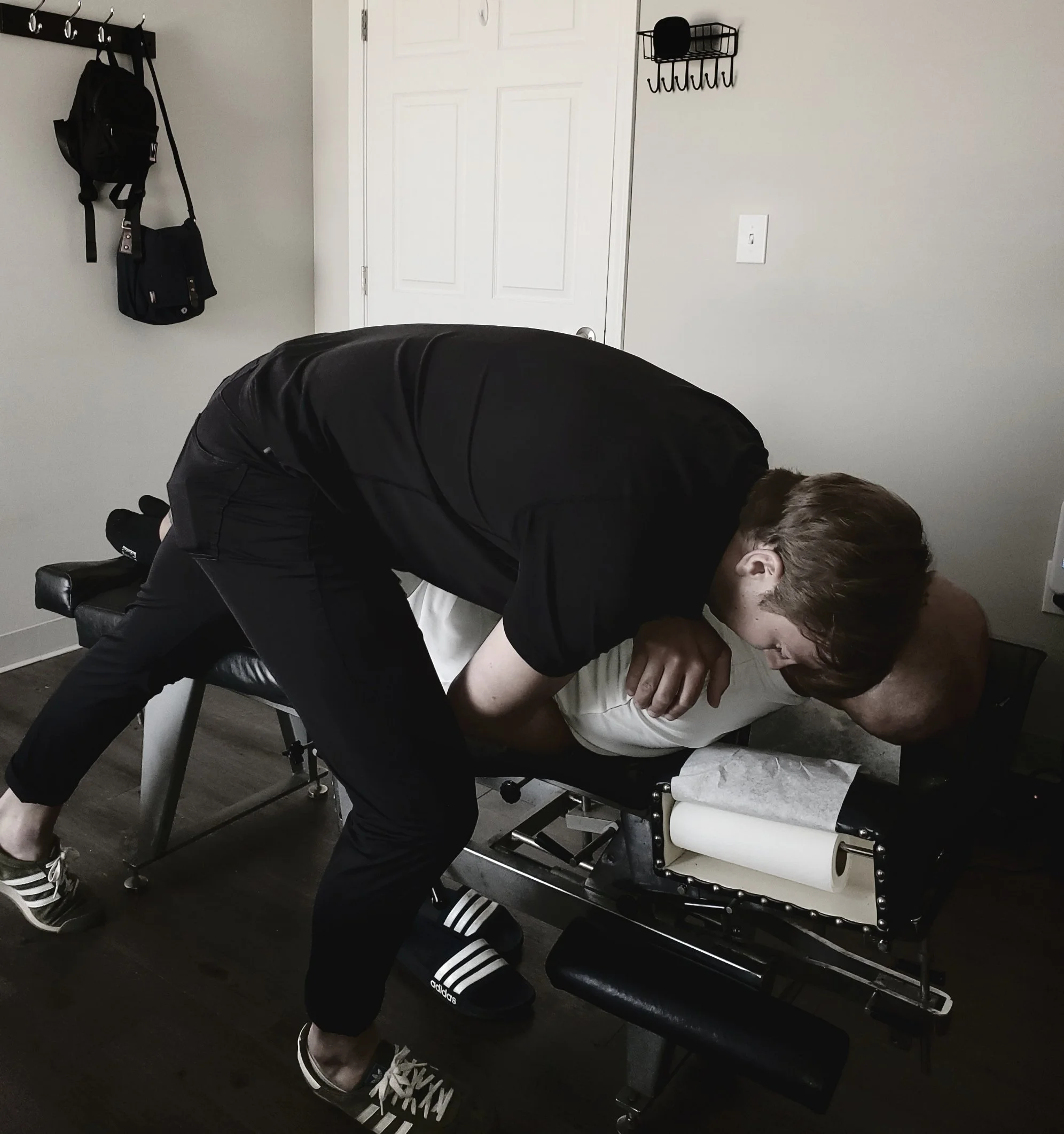 A person with light brown hair wearing a black jacket and black and white Adidas sneakers leaning over a hospital bed with someone lying on it, partially visible and covered with a white blanket. The room has gray walls, a white door, a black wall-mounted coat rack with bags, and a black wall-mounted hook organizer. The floor is dark wood.