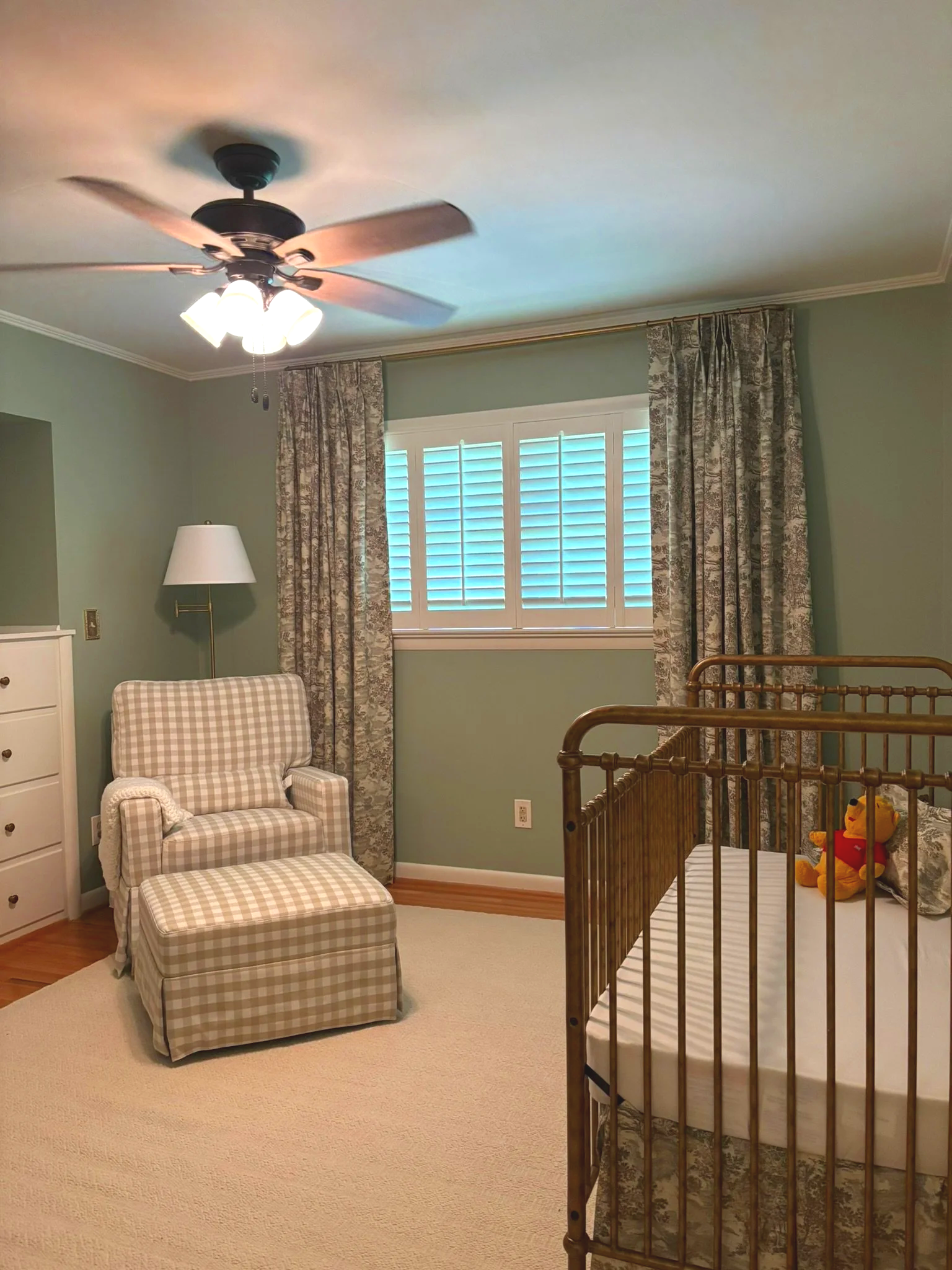 A bedroom with beige carpet, green walls, a window with blinds and floral curtains, a white armchair with a checkered slipcover and matching footstool, a white dresser, a brass crib with a stuffed Winnie the Pooh toy, and a ceiling fan with attached light fixtures.