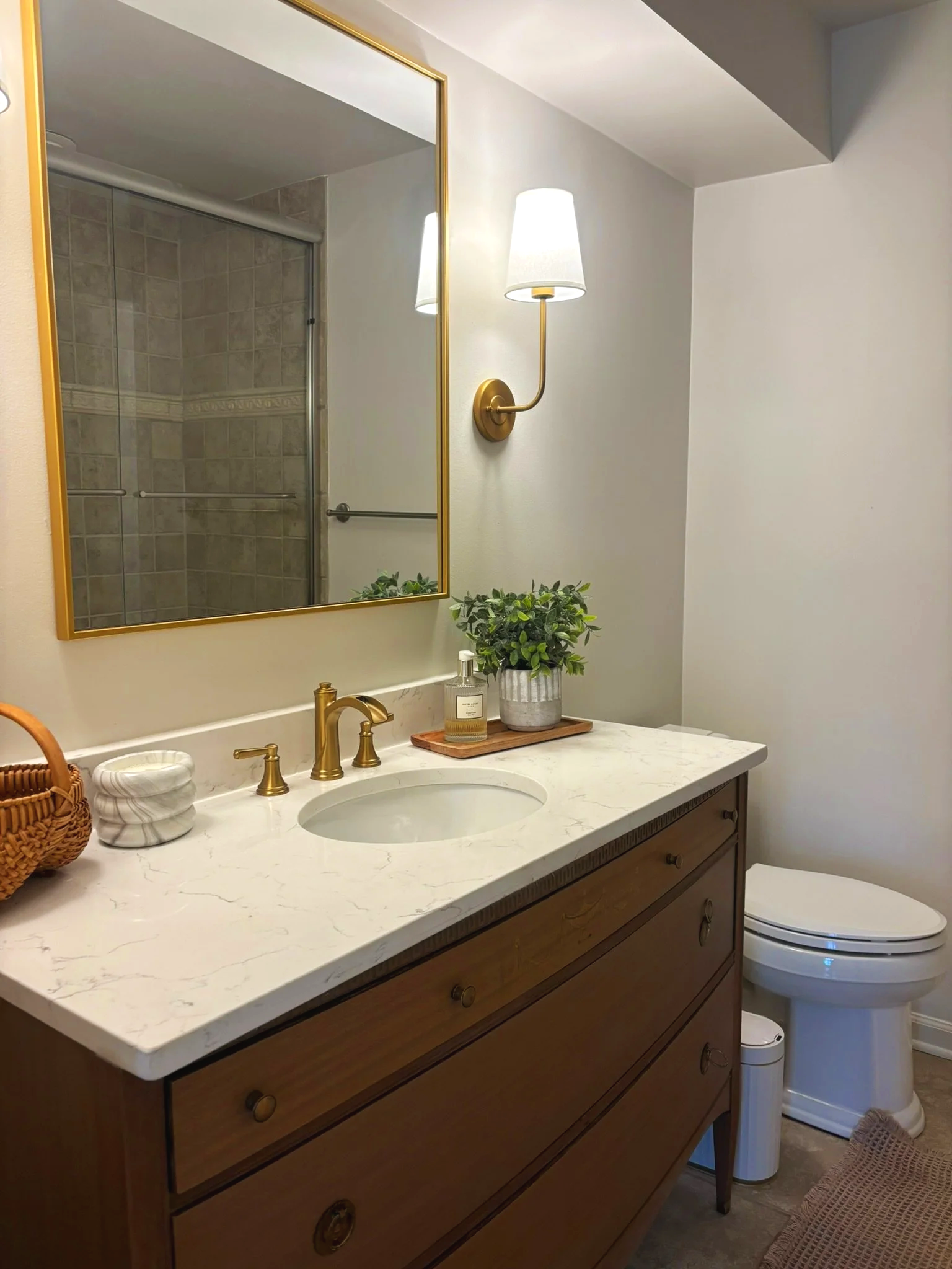 A bathroom vanity with a white marble countertop, a gold faucet, a potted plant, soap dispenser, and a bowl. A mirror with a gold frame hangs above, reflecting a shower with beige tiled walls. To the right, a white toilet with a woven cover is visible along with a trash bin and a small brown mat.