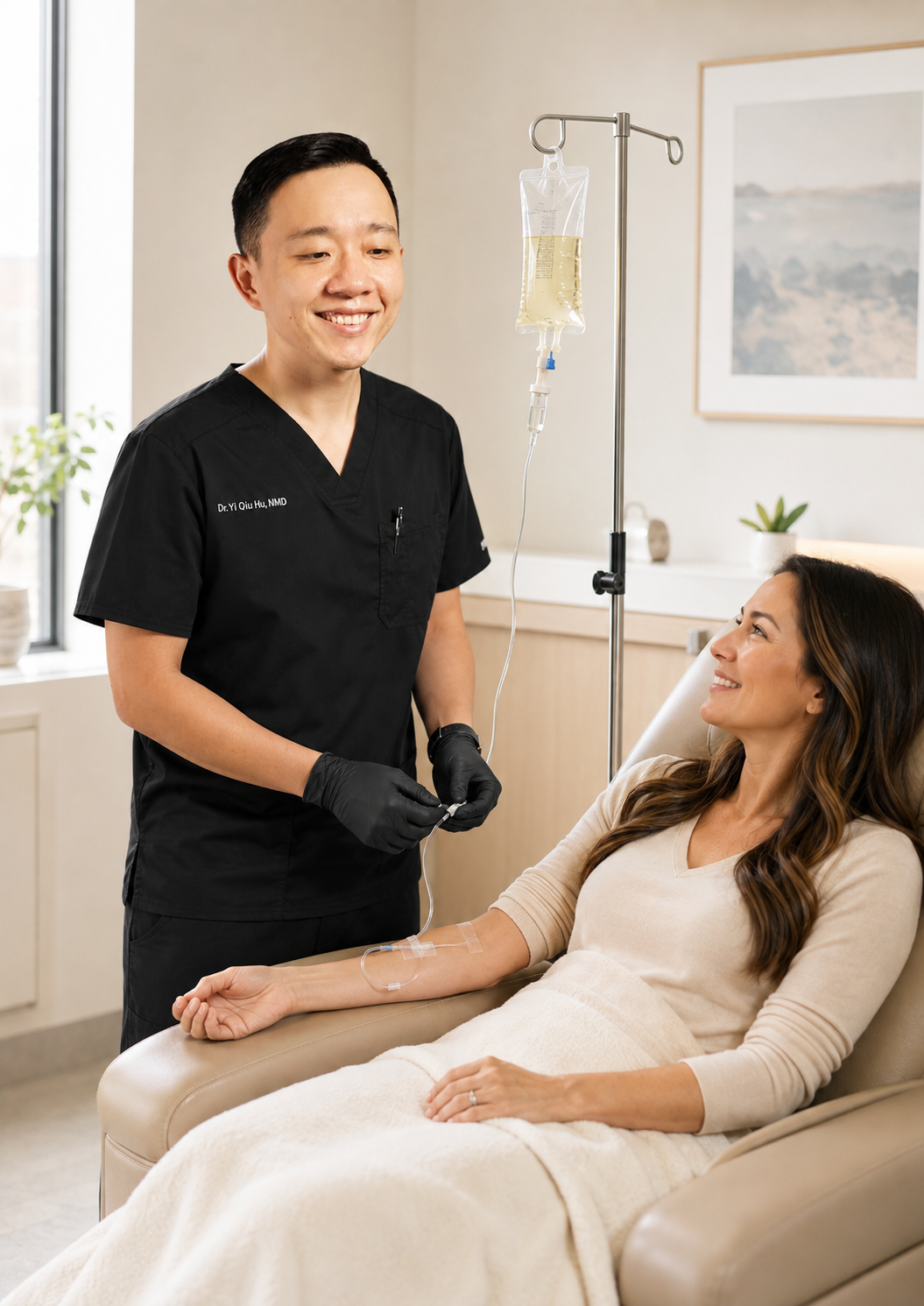 A male healthcare professional in black scrubs and black gloves stands next to a woman sitting in a hospital bed. The woman has an IV in her arm and is smiling at the healthcare worker. The setting is a bright hospital room with a window and a piece of artwork on the wall.