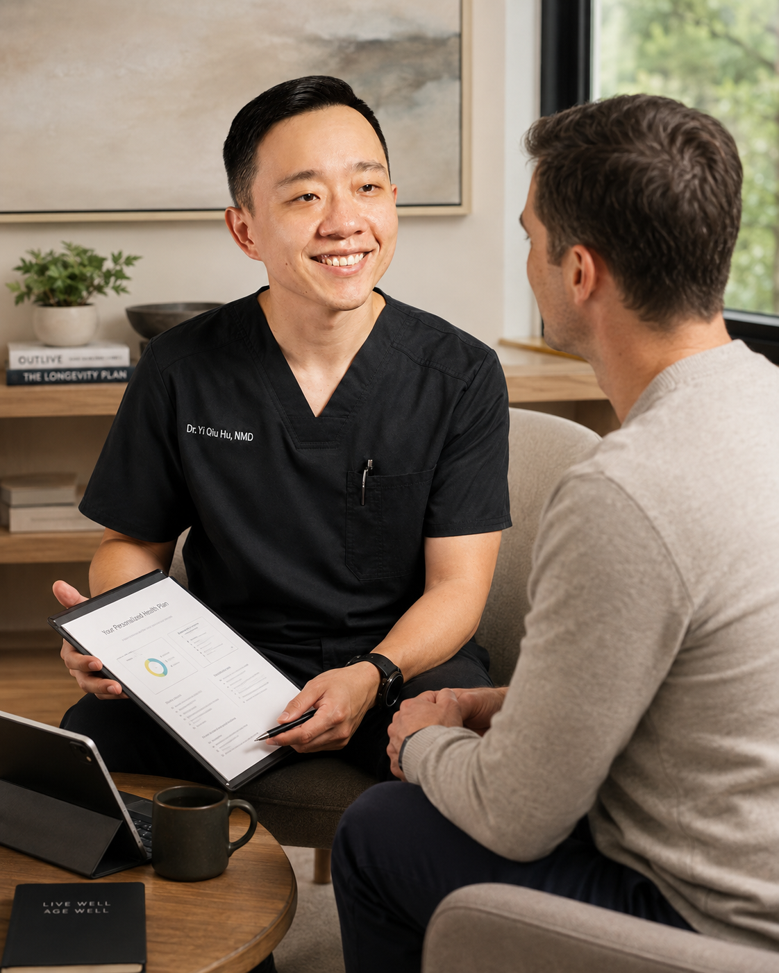 A healthcare professional wearing black scrubs talks with a patient in a consultation room. The healthcare worker holds a tablet displaying a health plan, and both are engaged in a conversation.