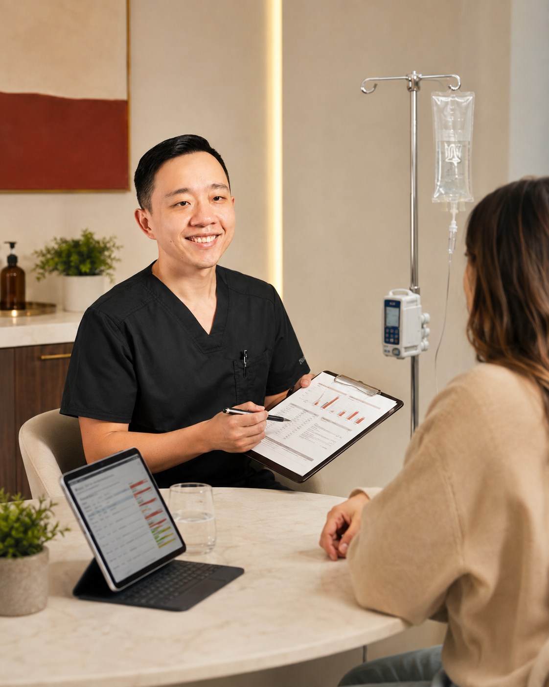 A healthcare professional, wearing black scrubs, smiling while holding a clipboard, sitting at a table with a patient in a beige sweater. Medical equipment, including an IV drip, is visible in the background.