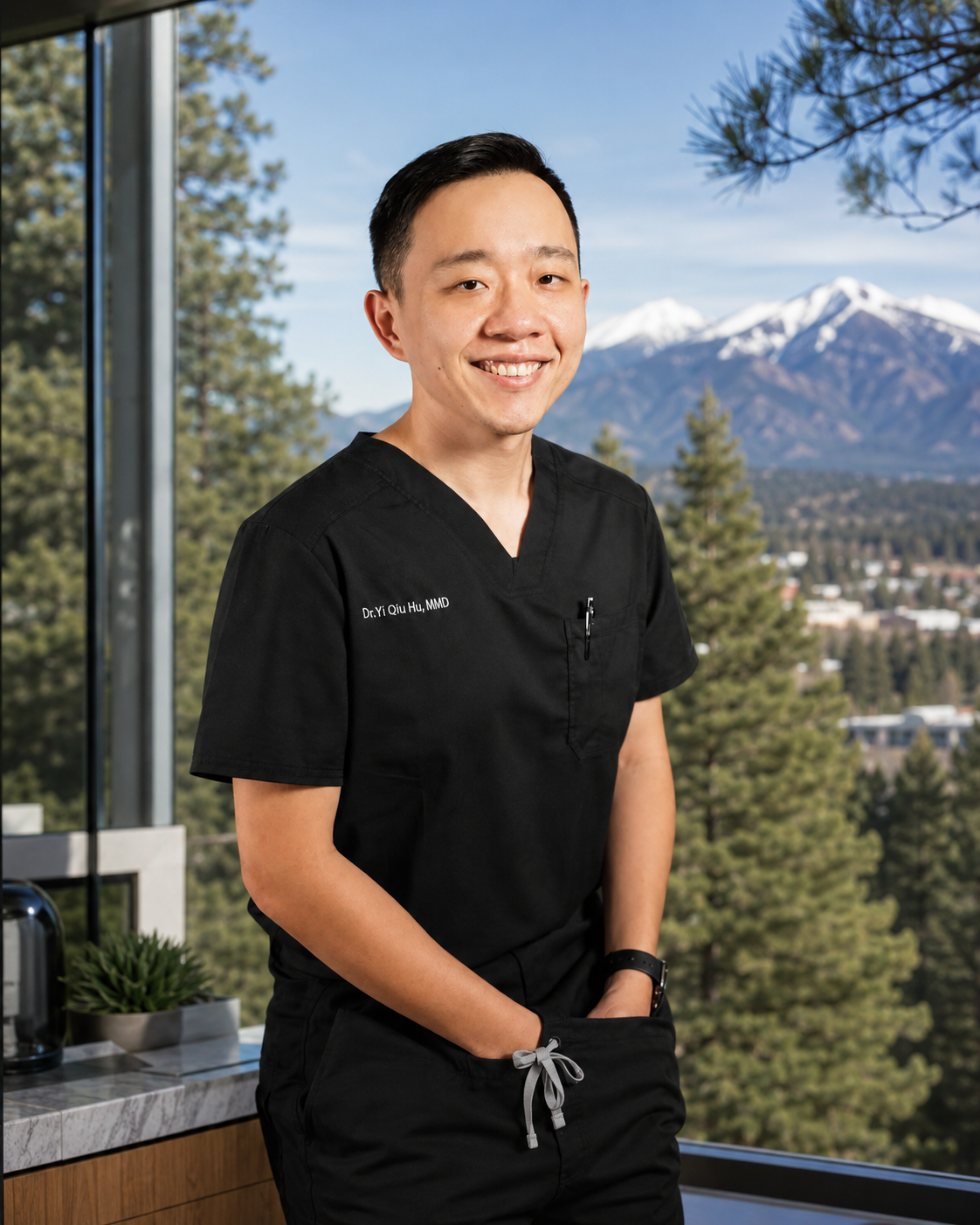 A young man in black medical scrubs with a mountain and forest background, smiling at the camera.