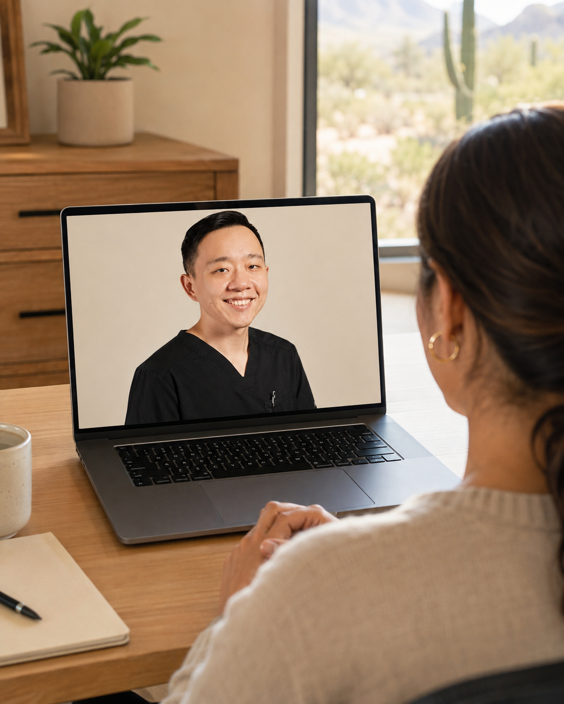 A woman on a video call with a man in medical scrubs on her laptop at a desk in a bright room with a window showing desert landscape with cacti