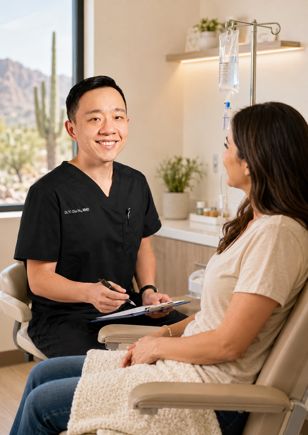 A smiling male healthcare professional speaking with a female patient in a medical office, with an IV drip and desert landscape visible outside the window.