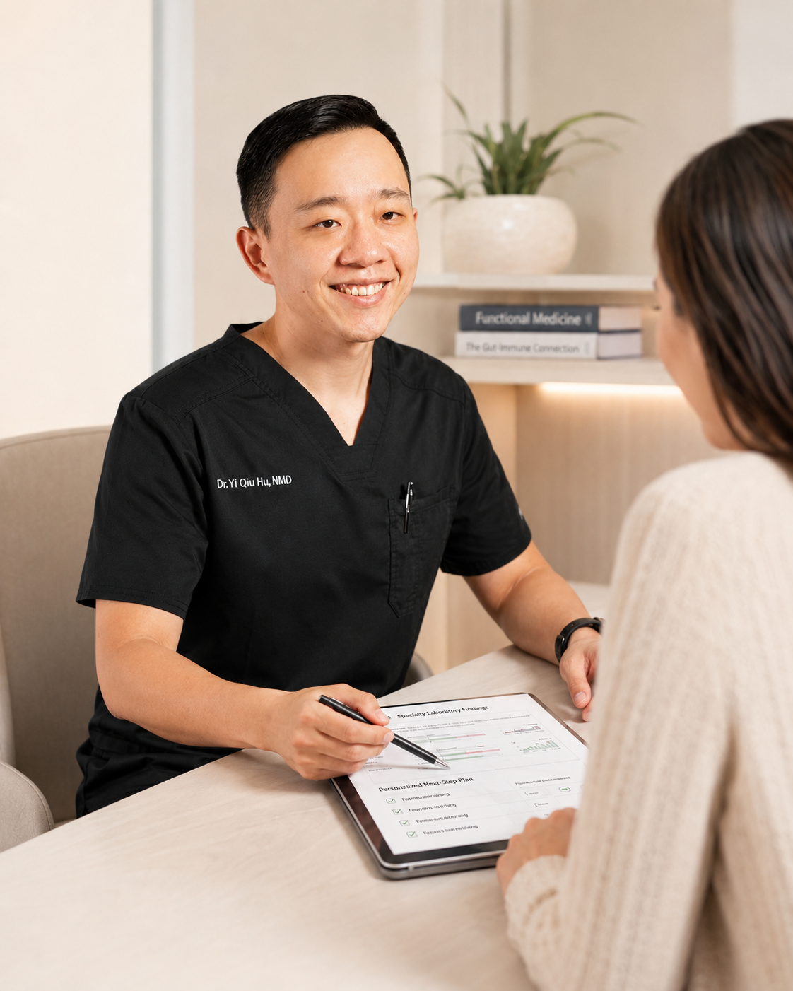 A male healthcare professional with short black hair, wearing black scrubs, smiling and sitting at a desk with a woman, holding a pen over a health plan document on a tablet in a medical office.