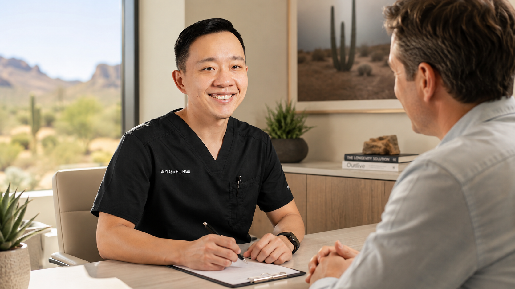 A smiling doctor in black scrubs talking to a patient in an office setting with desert landscape outside the window.