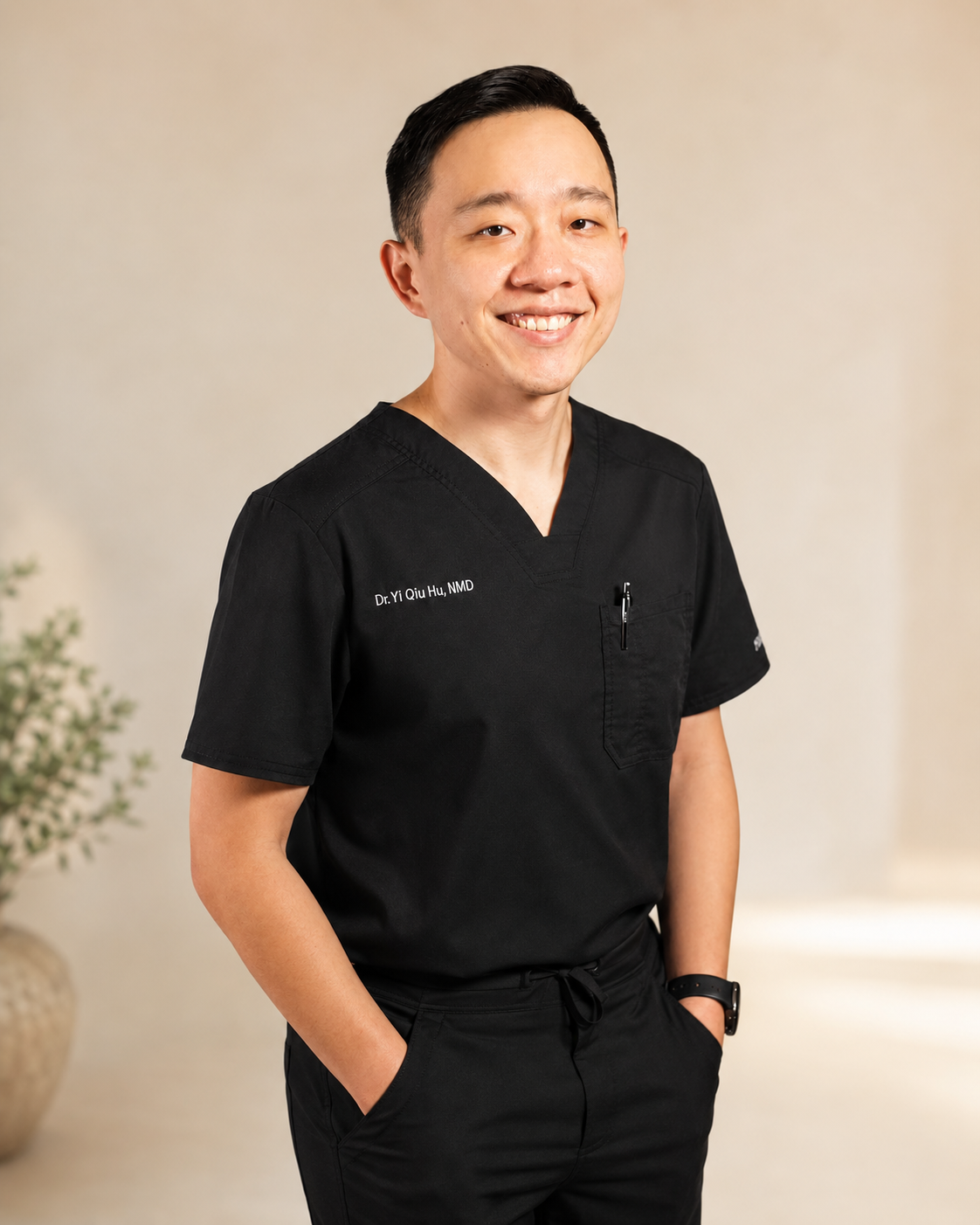 A smiling man in black medical scrubs posing indoors with hands in pockets, a plant in the background.