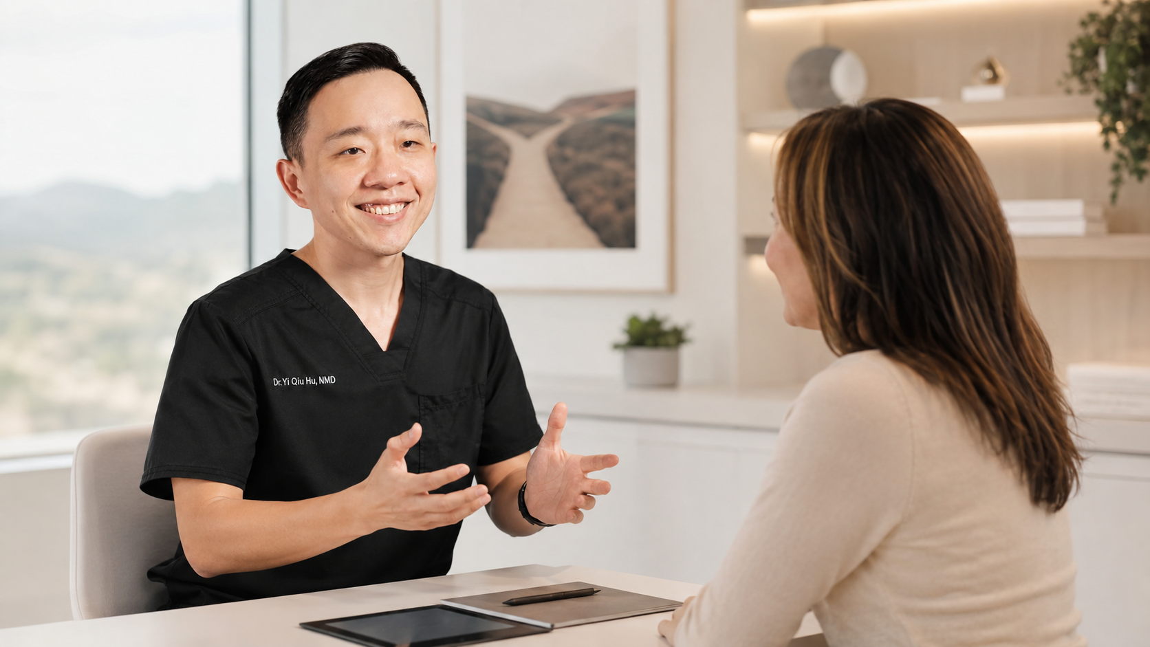 A smiling male doctor in black scrubs talking with a female patient in an office setting, with a tablet and pen on the table.