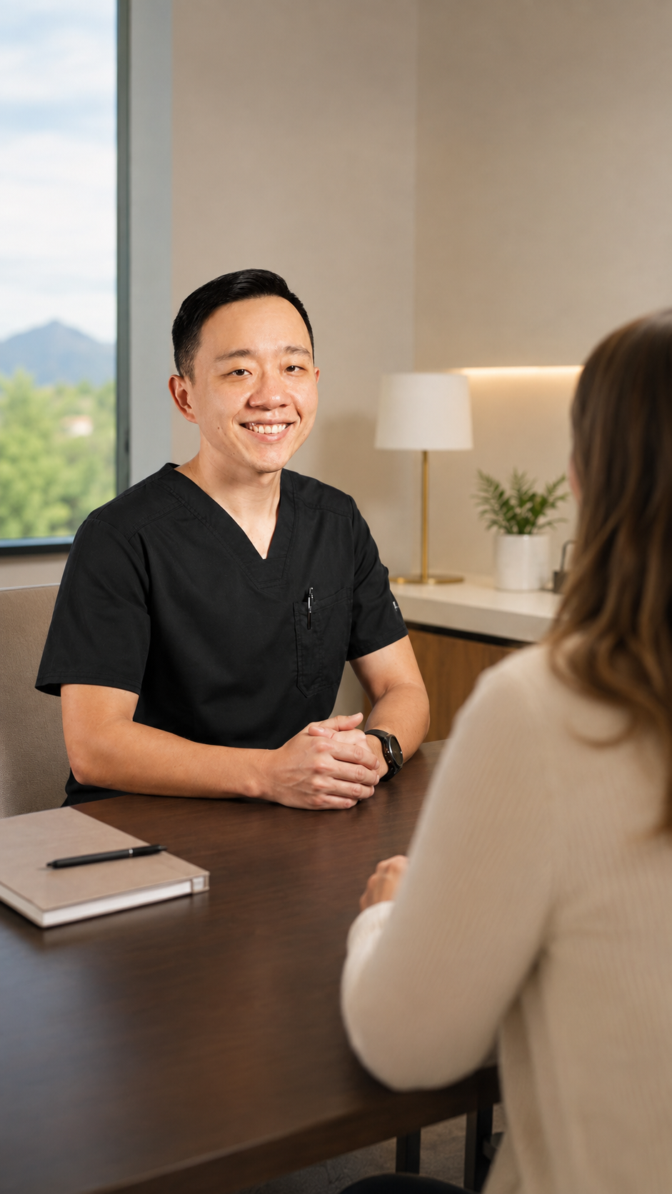A healthcare professional in black scrubs smiling and talking to a woman in a cream sweater during an appointment in a well-lit office with a large window, a table, a notebook, and a potted plant.
