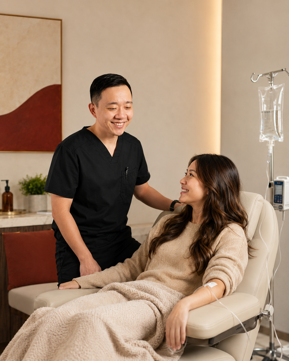 A male healthcare professional in black scrubs smiling and talking to a female patient sitting in a comfortable chair with an IV drip in a modern medical office.