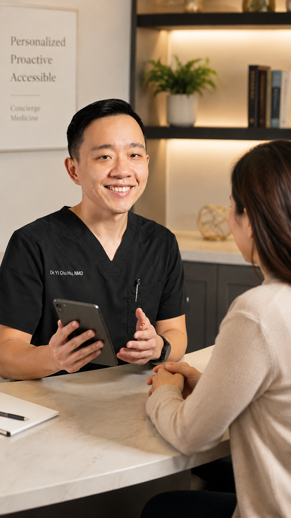 A doctor in black scrubs smiling and holding a smartphone while talking with a patient seated across from him in a reception area or consultation room.