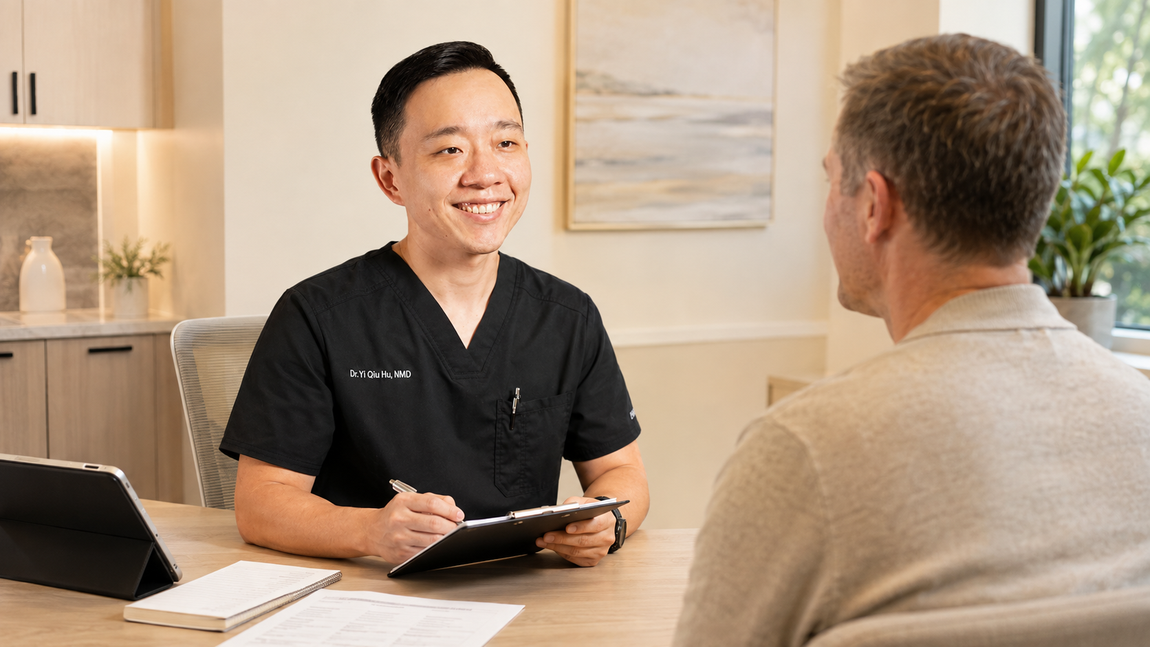A doctor in black scrubs smiling and holding a clipboard while talking to a male patient in a beige shirt in a medical office.