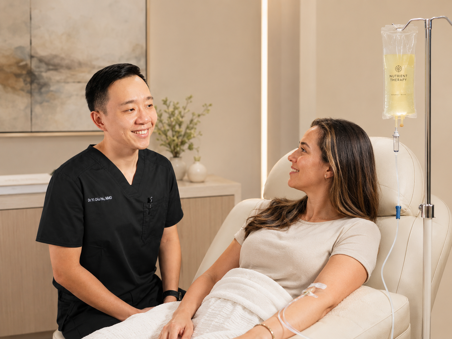 A female patient is lying in a hospital bed with an IV drip, smiling and talking to a smiling male healthcare professional in black scrubs, in a medical facility.