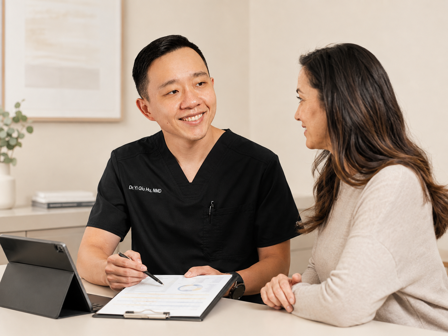 A doctor in a black medical uniform with the name 'Dr. Yi Qiu Hu, NMD' talking to a woman during a consultation. They are sitting at a desk with a tablet and a clipboard, having a friendly discussion in a medical office.
