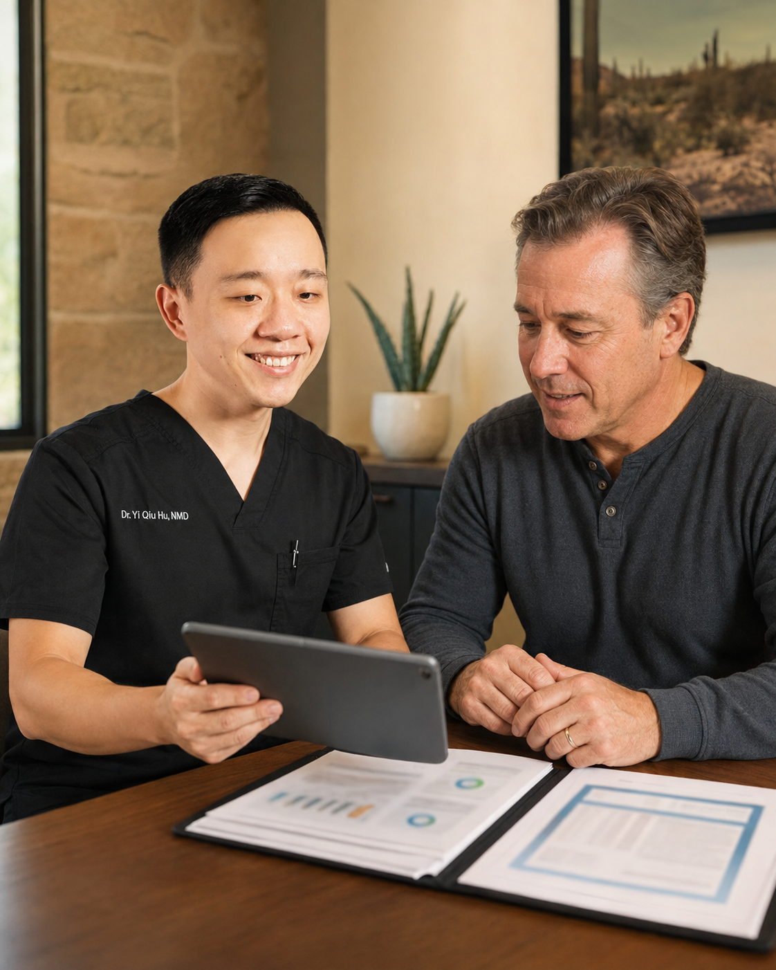 A healthcare professional showing something on a tablet to a middle-aged man in an office setting with a framed landscape photo and a potted plant in the background.