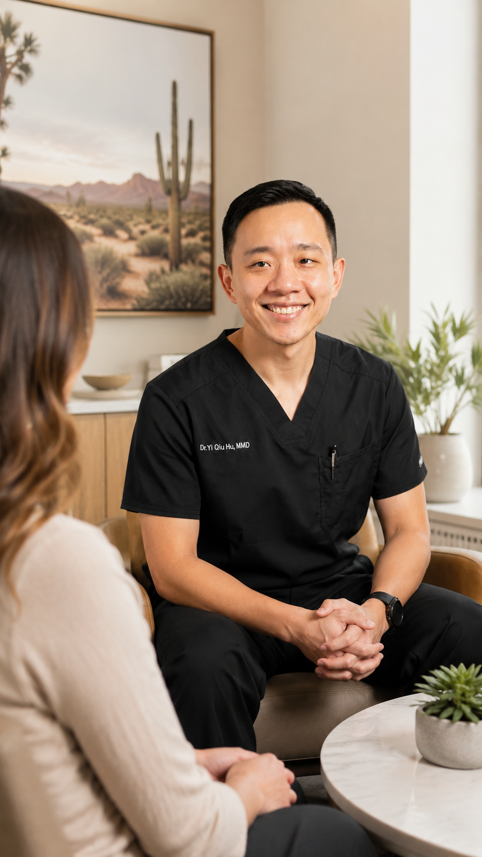 A smiling male doctor in black scrubs talking to a woman in a beige sweater in a cozy room with desert landscape art and plants.