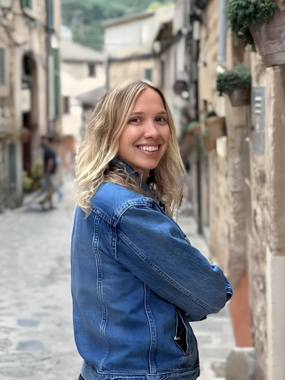 A young woman with blonde hair smiling while standing in a European-style cobblestone street, wearing a blue denim jacket.