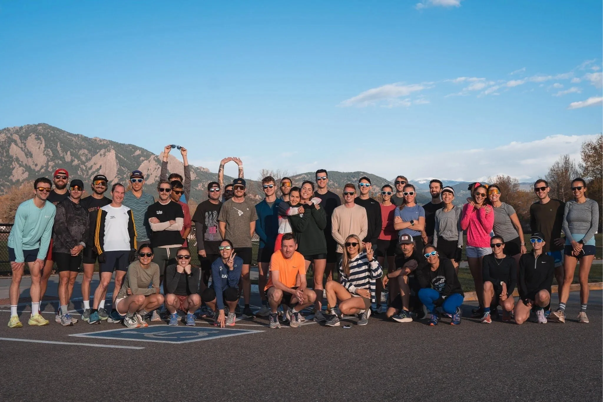 A large group of people gathered outdoors on a sunny day with mountain scenery in the background, some wearing athletic clothing and sunglasses, smiling and posing for a group photo.
