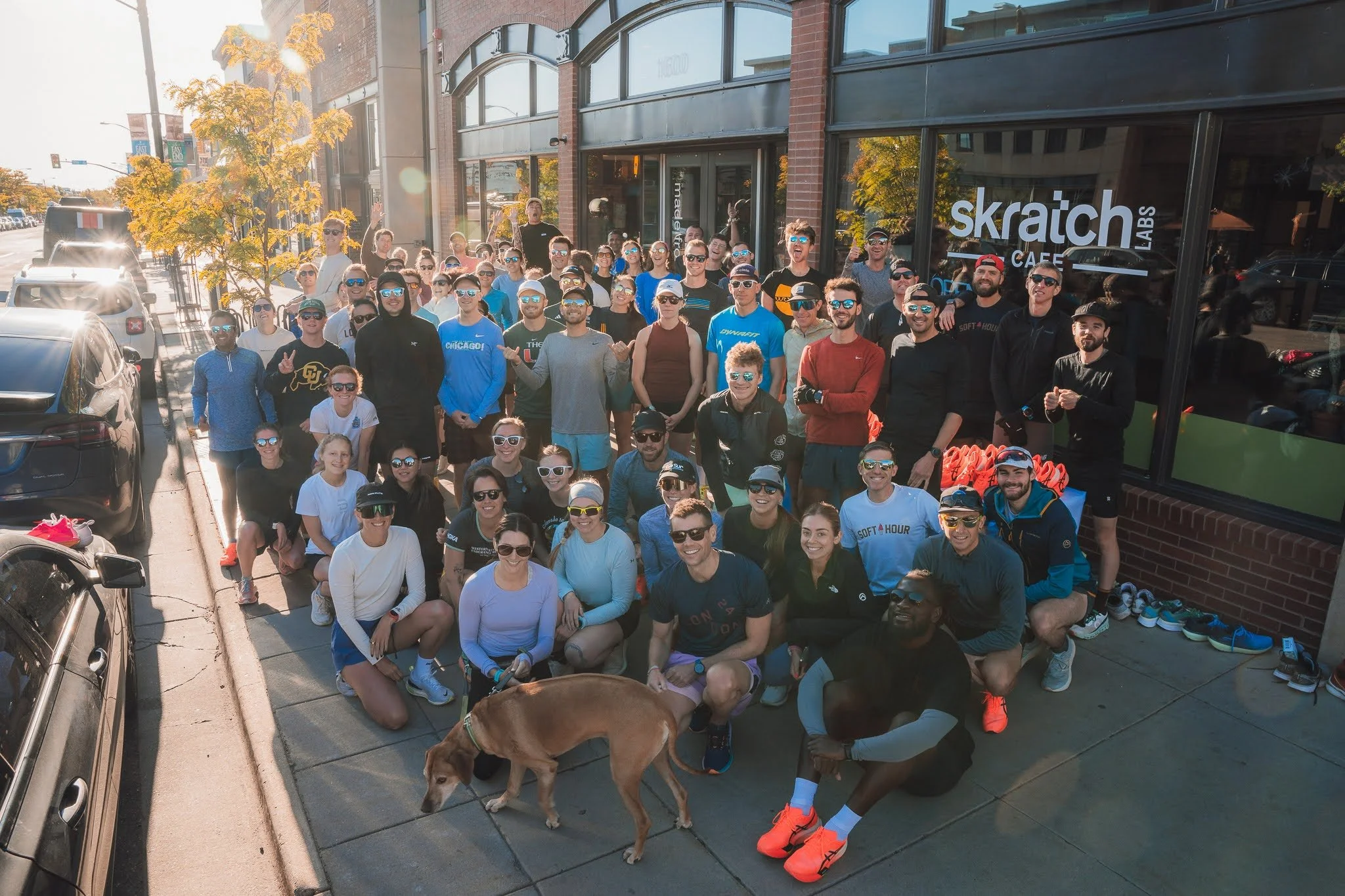 A large group of people standing outside on a sunny day in front of a building with the sign "skraitch CAFE". Many are wearing sunglasses and athletic clothing, some are smiling and waving, and a dog is visible in the foreground.