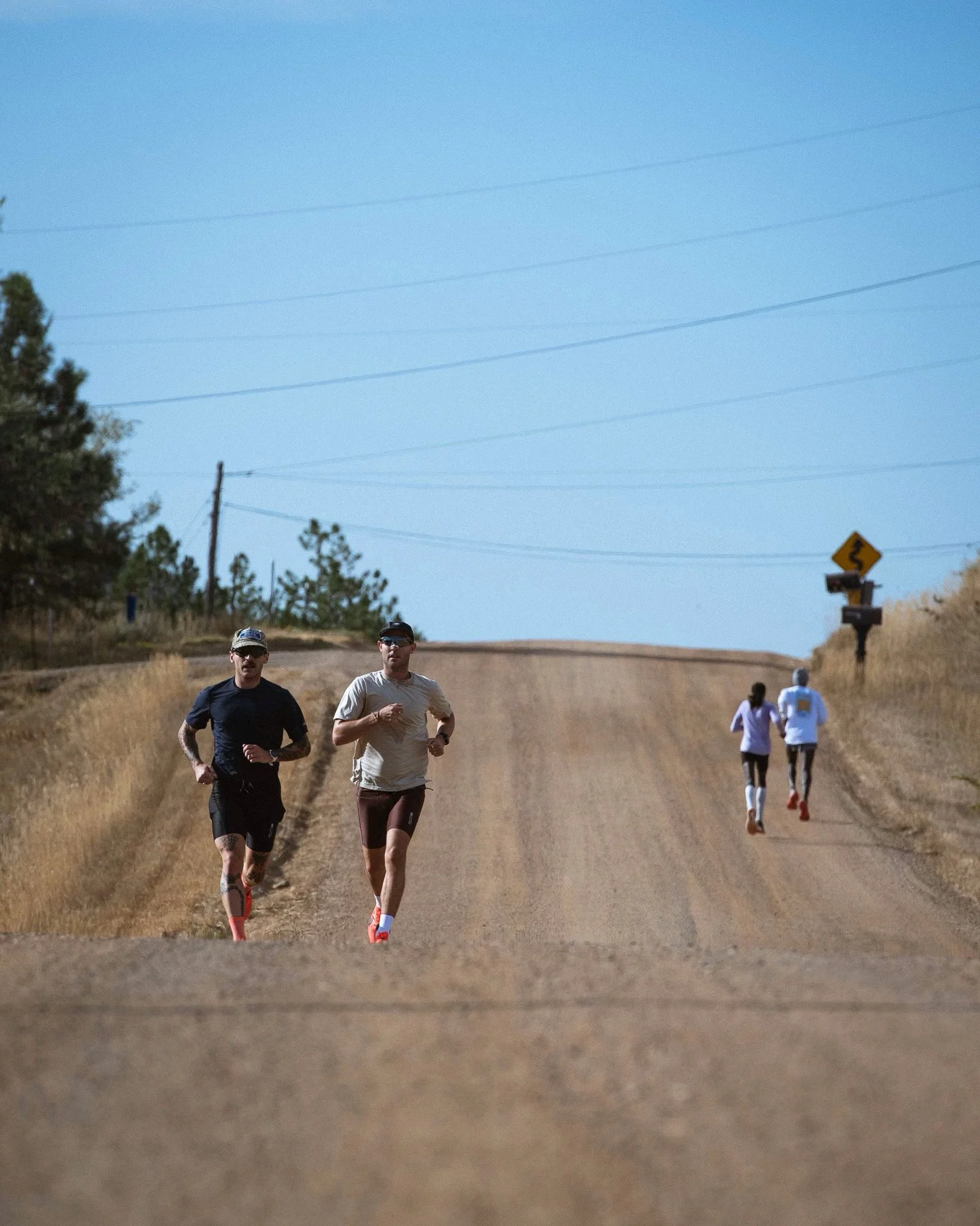 A group of people running on a dirt road through a rural landscape with dry grass and trees, some wearing athletic clothing and shoes.