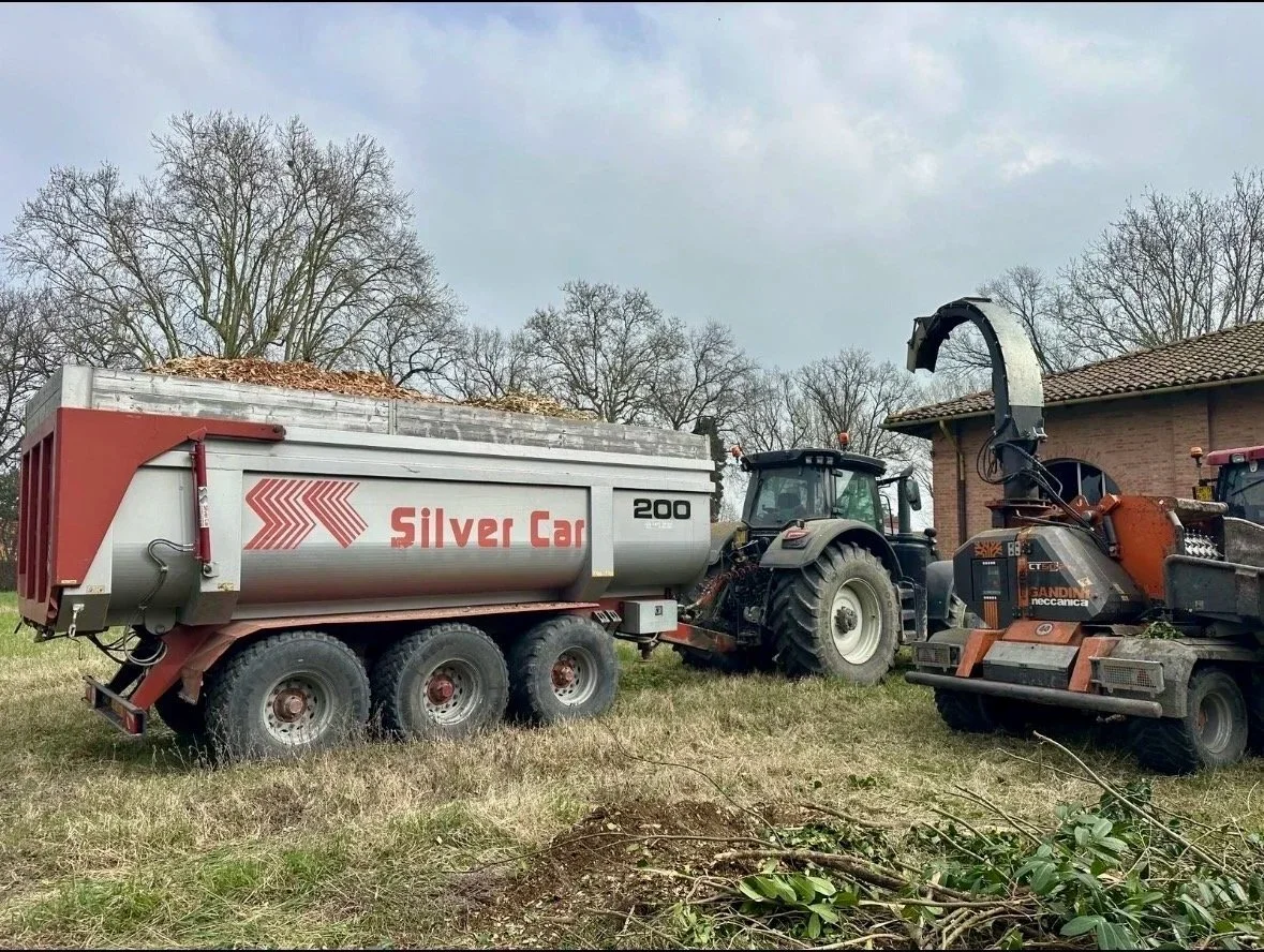 Macchine agricole in un campo: una trattrice con rimorchio carico di foglie e detriti, una tritatrice con un trasportatore a bocca, e un edificio in background, sotto un cielo nuvoloso.
