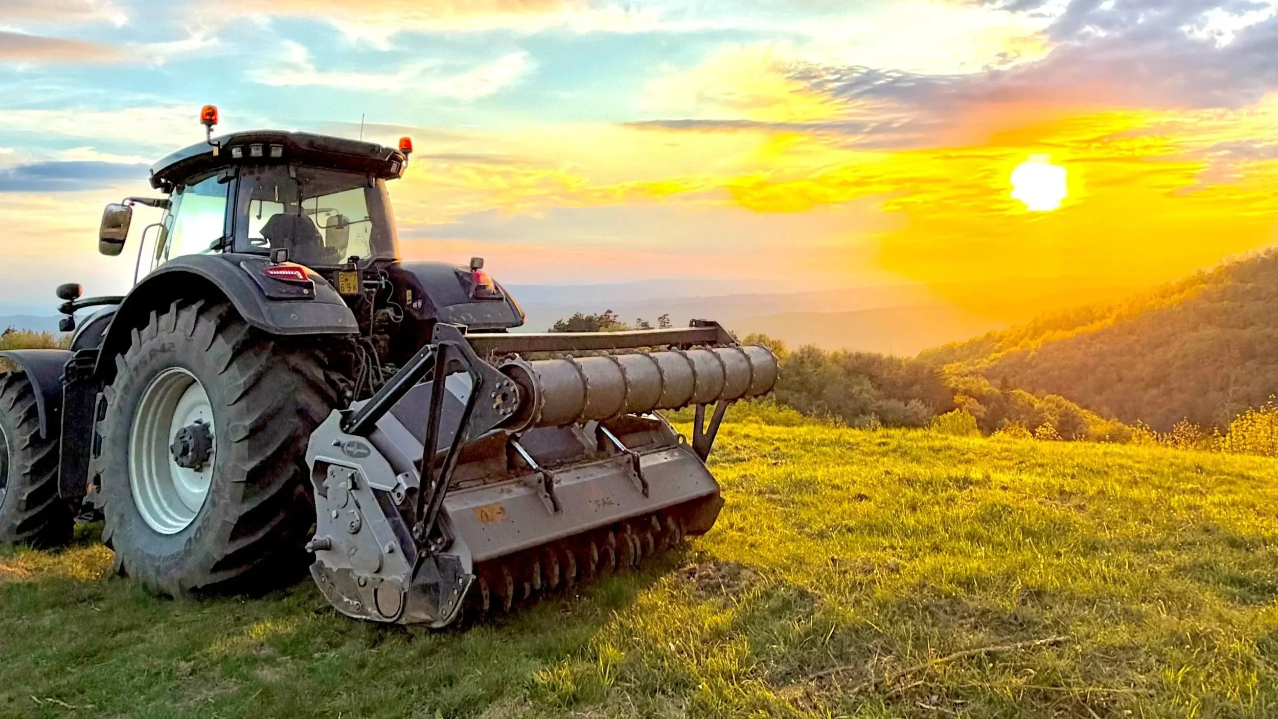 Un trattore in un campo durante il tramonto, con il sole basso sull'orizzonte e colline verdi sullo sfondo.